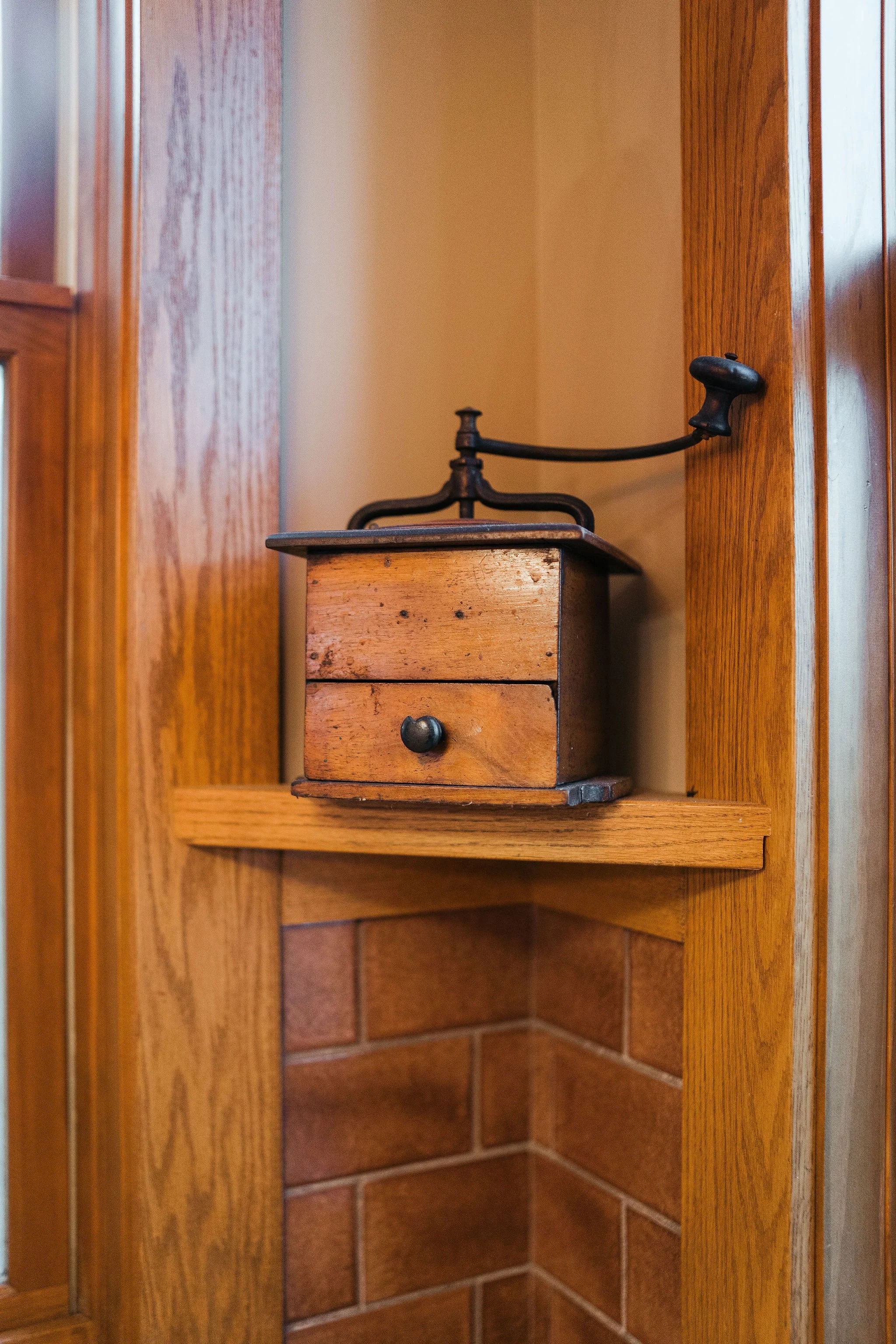Old wooden hand-crank coffee grinder on a shelf in a kitchen with brick and wood paneling.