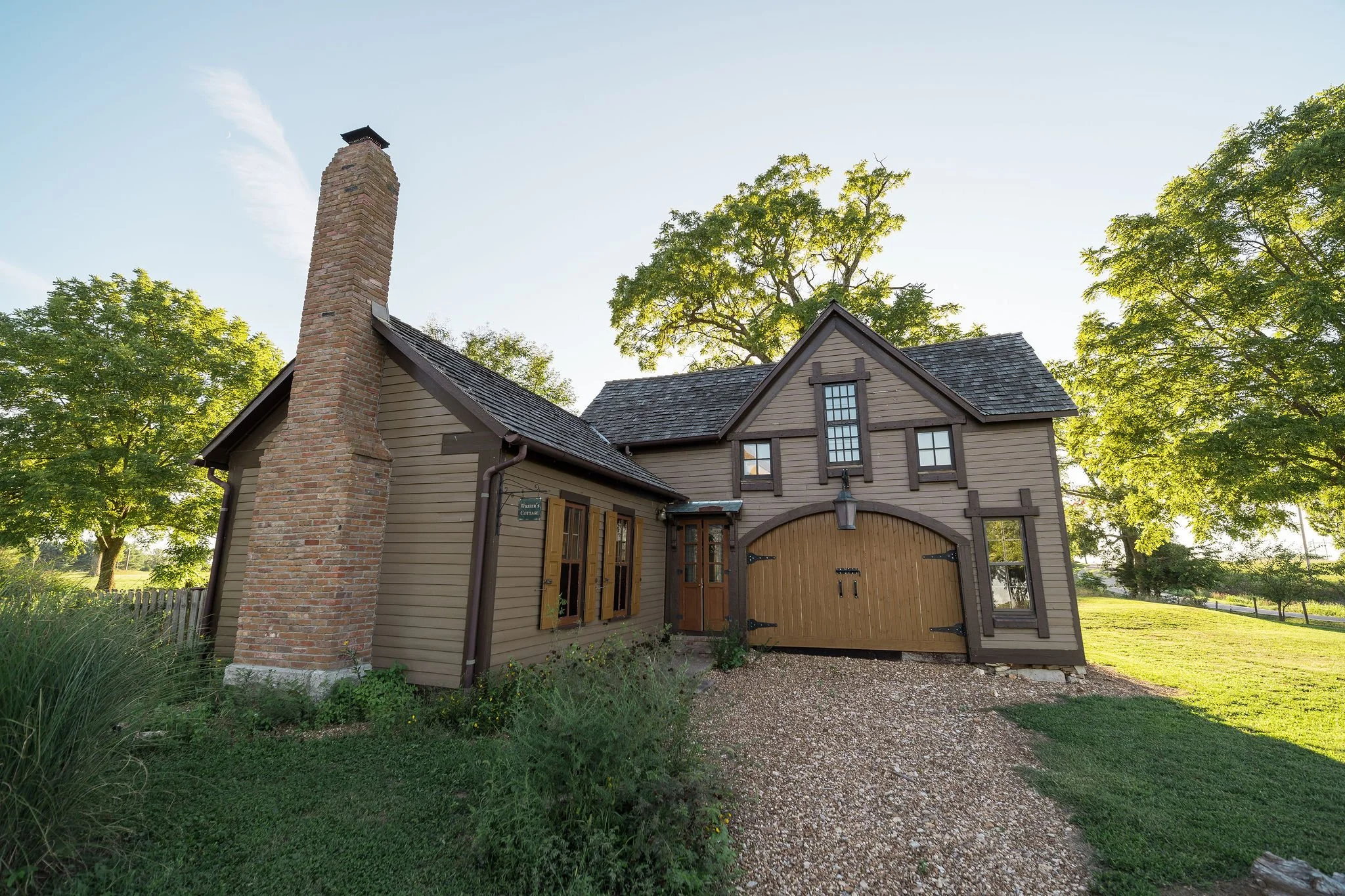 A cozy, rustic house with a brick chimney, wooden garage door, and multiple windows, surrounded by green trees and a gravel pathway.