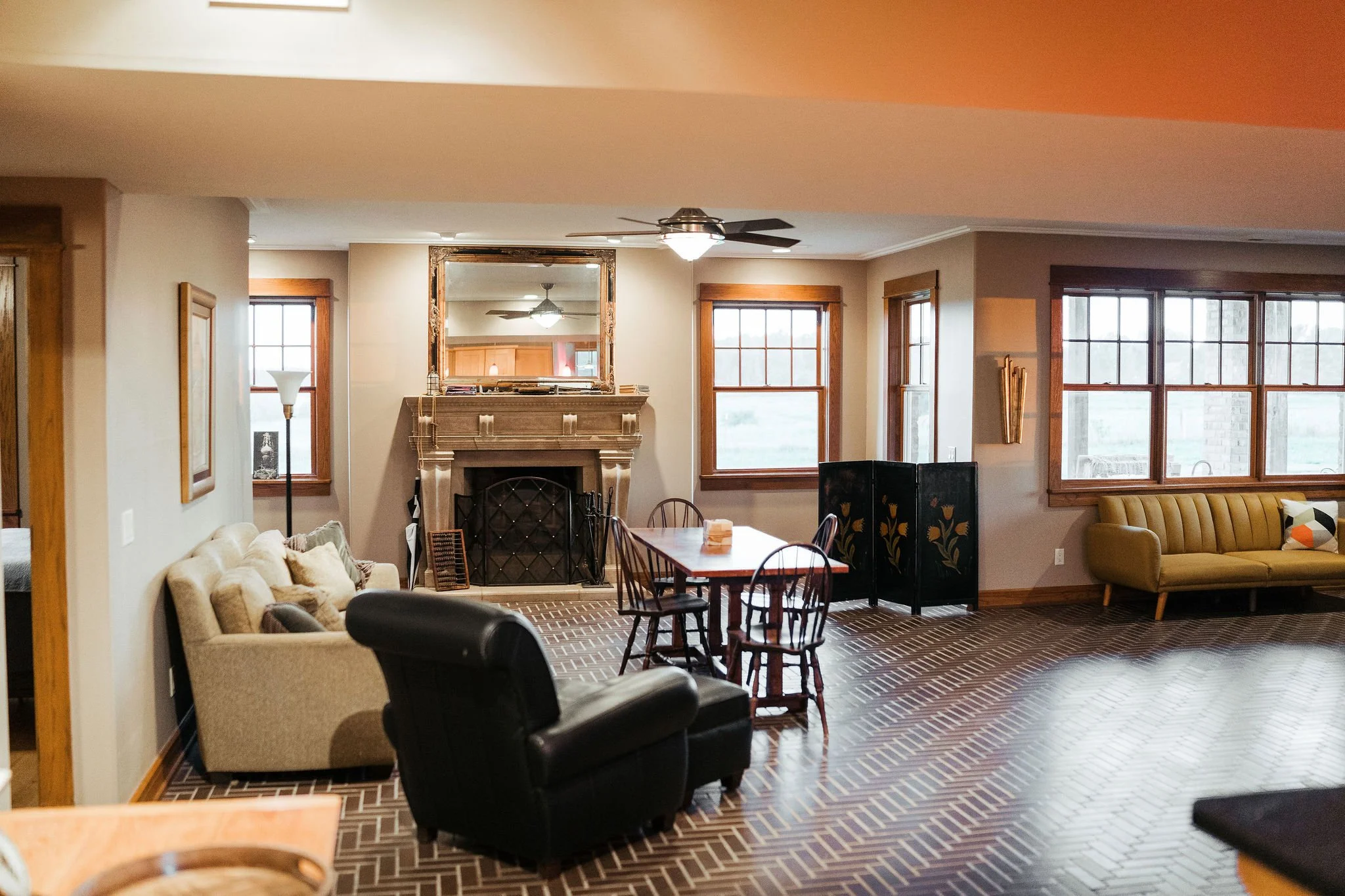 Living room with a brick fireplace, wooden window frames, a beige sofa, a black armchair, a dining table with chairs, and a yellow sofa, with large windows letting in natural light.