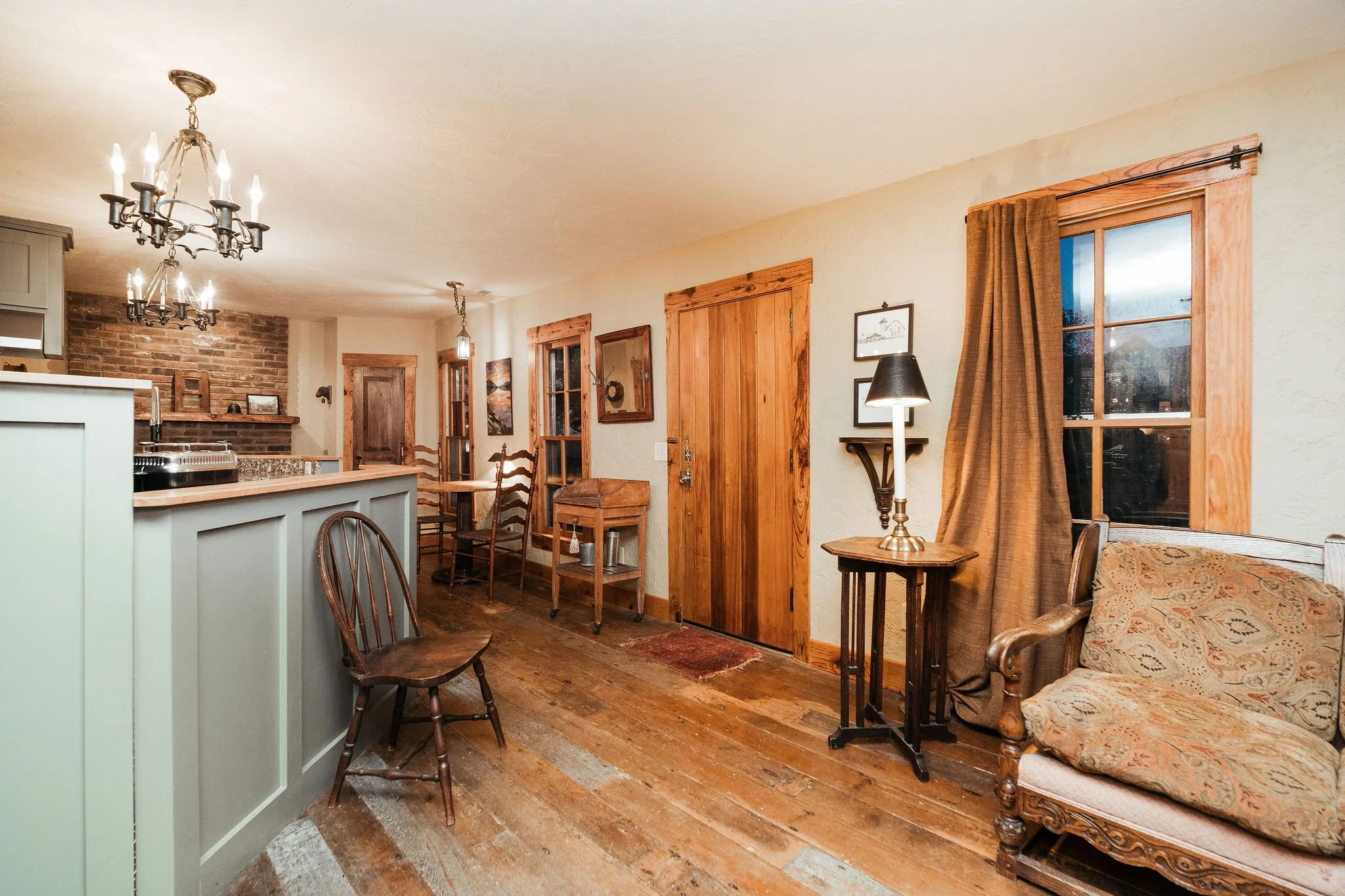 Living room with wooden floors, a window with curtains, a wooden armchair with fabric cushion, side table with lamp, and a kitchen counter with hanging chandeliers.