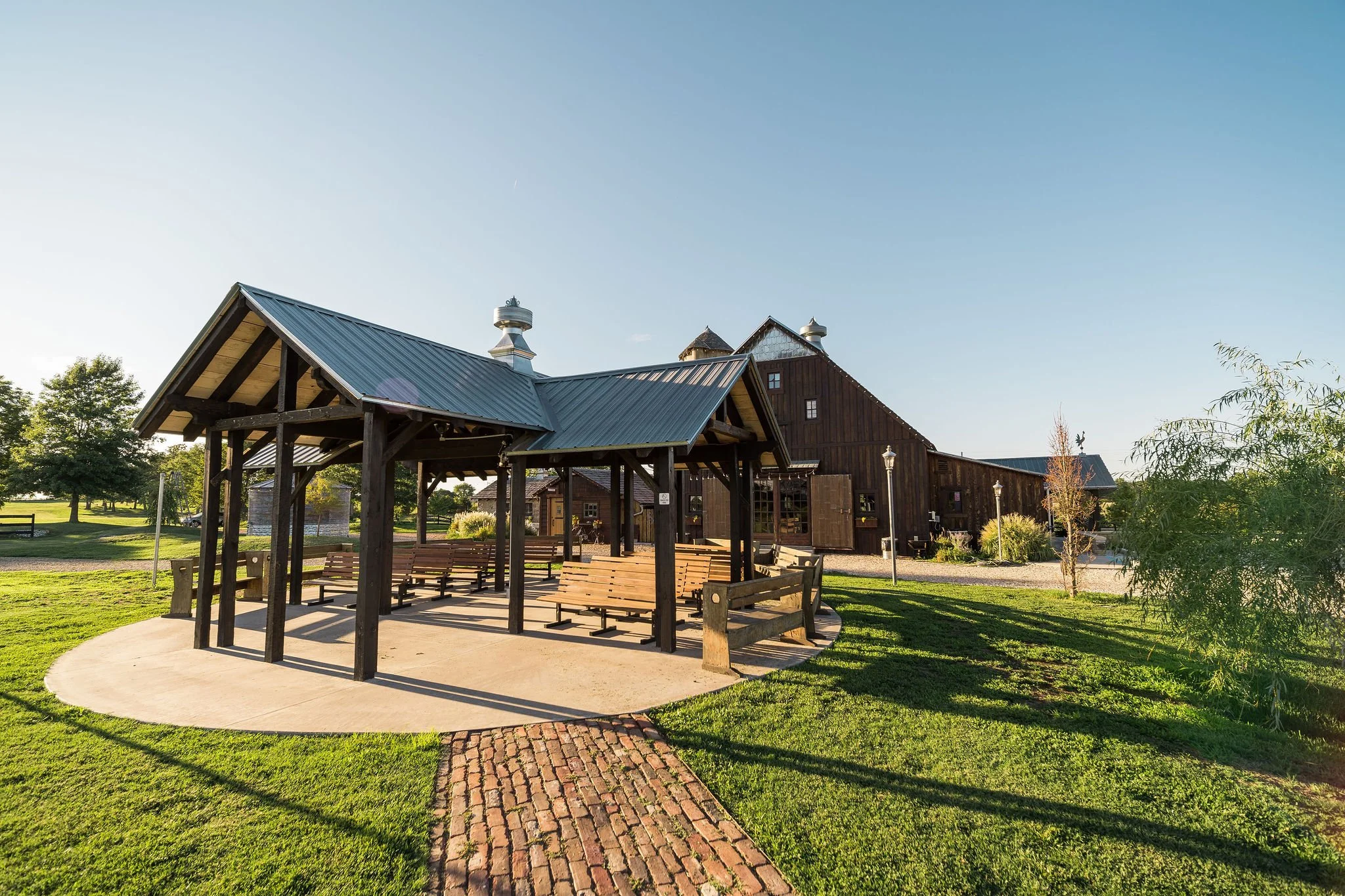 A rustic outdoor pavilion with wooden benches and a metal roof in a grassy park, with a large wooden barn-style building in the background and a clear blue sky.