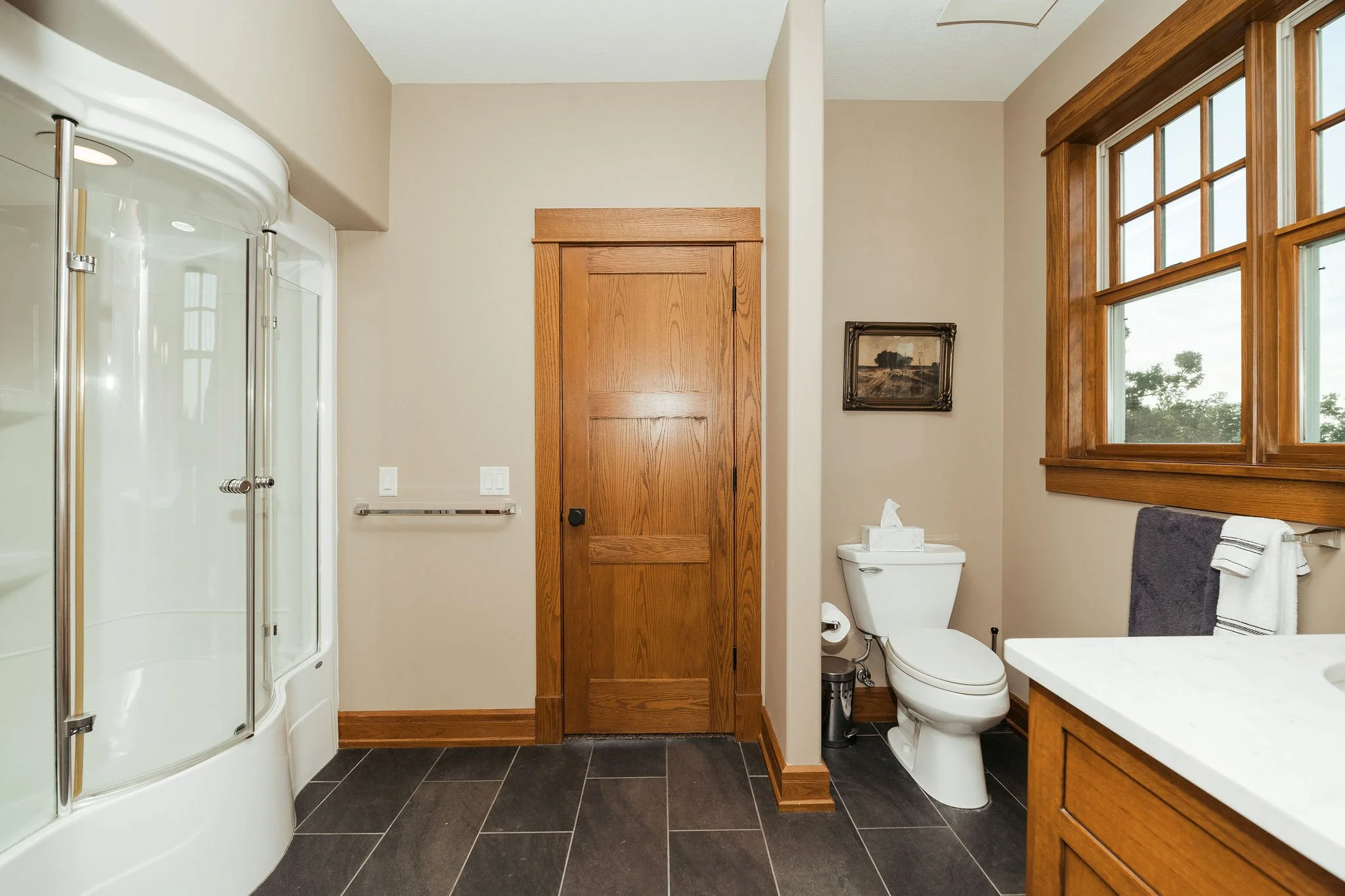 Bathroom with wooden door, large window with wooden frame, built-in shower cabin, toilet with tissue box, framed landscape painting, and wooden vanity with a white countertop.