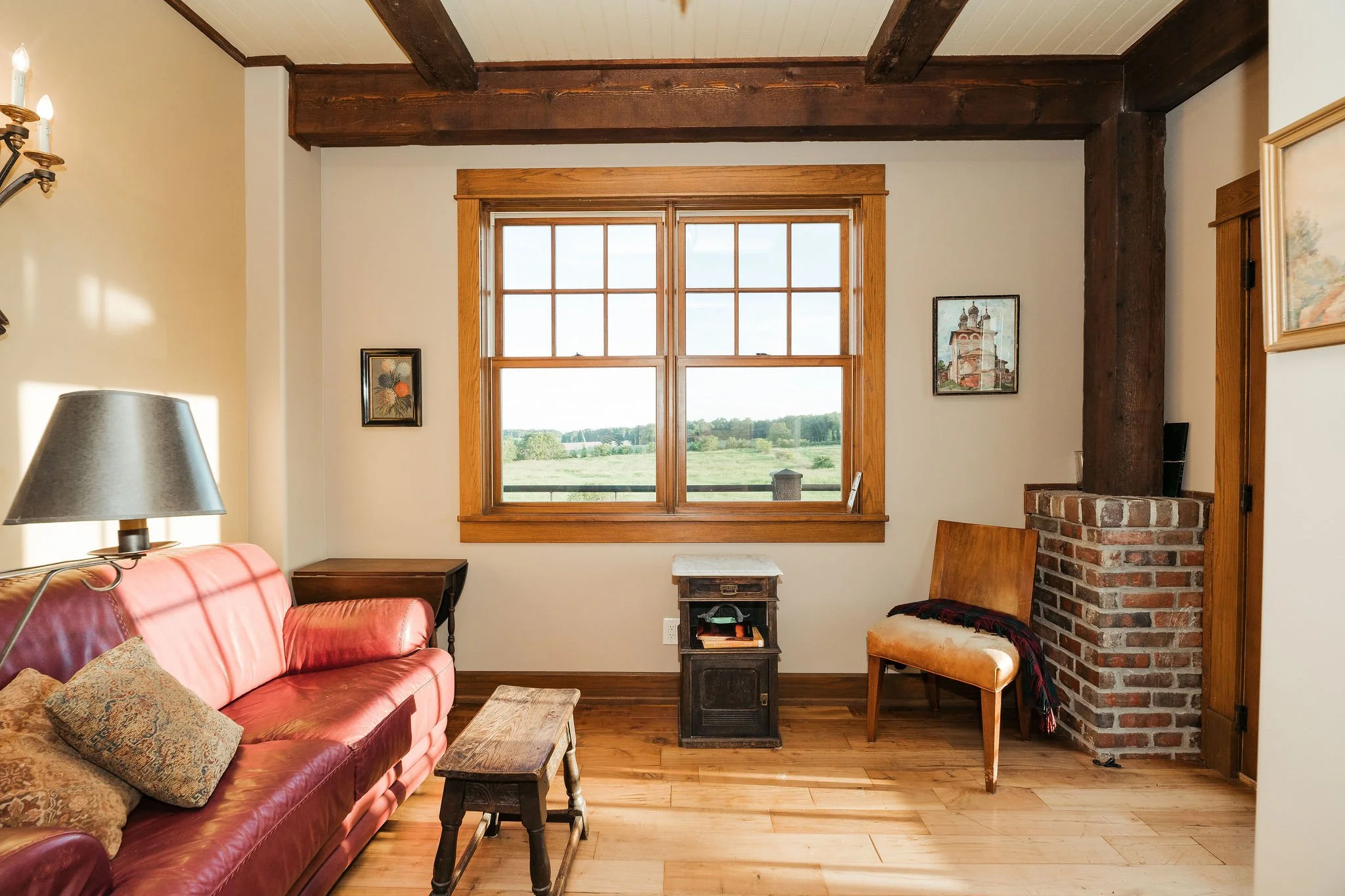 A cozy living room with a large window showing a green landscape outside. Contains a red leather sofa with decorative pillows, a wooden bench, a small side table, a wooden chair with a blanket, and a brick chimney with a wooden beam. The room has woo