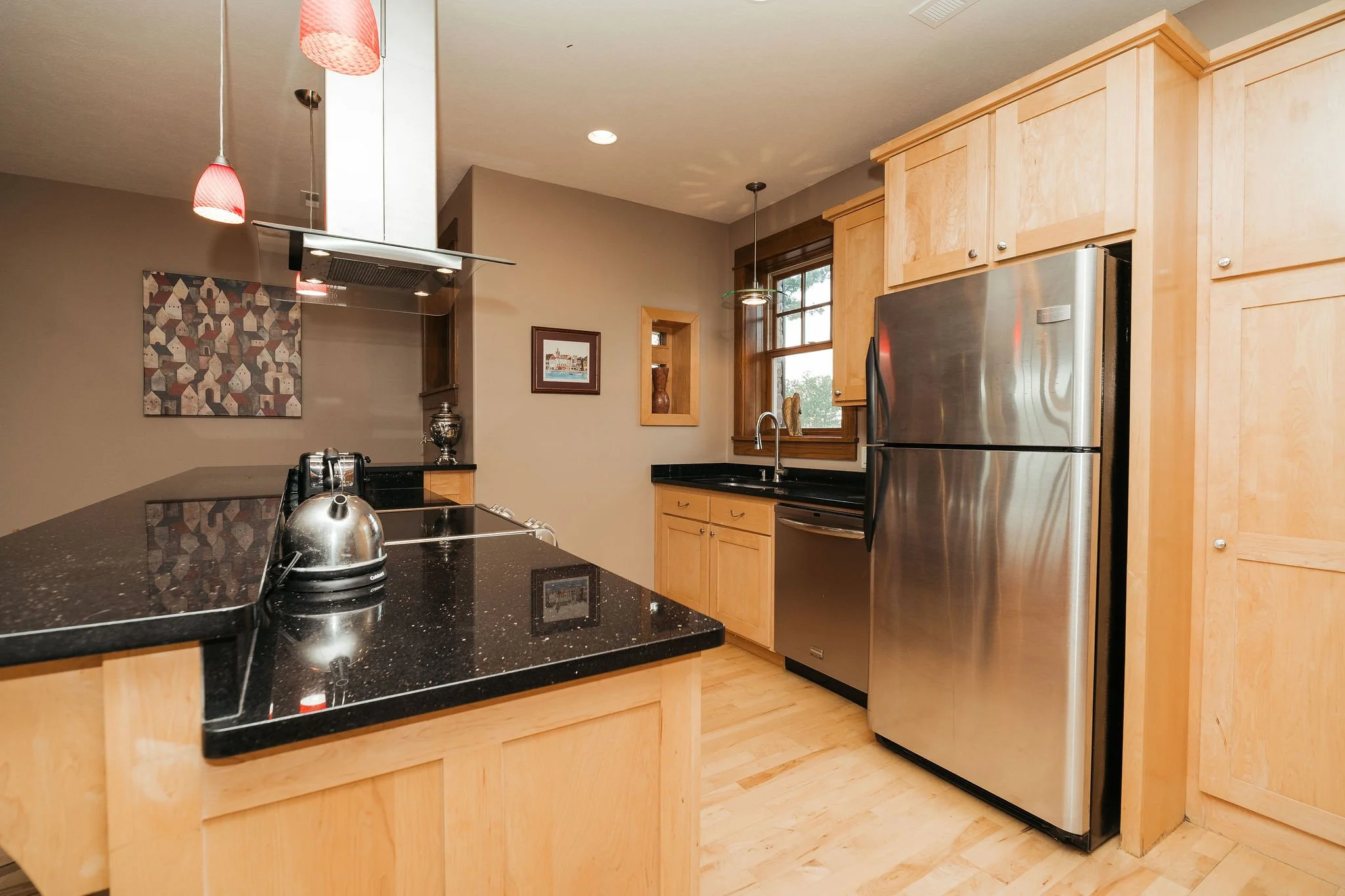 Modern kitchen with light wood cabinets, black countertops, stainless steel refrigerator, kitchen sink under window, and decorative wall art