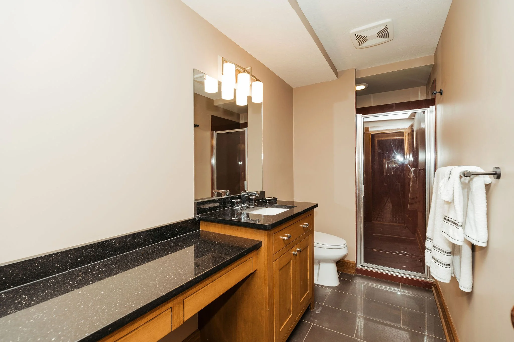 Bathroom with beige walls, black granite countertop, wooden vanity, mirror, modern light fixture, white toilet, glass shower with dark brown tiles, and a towel rack with white towels.