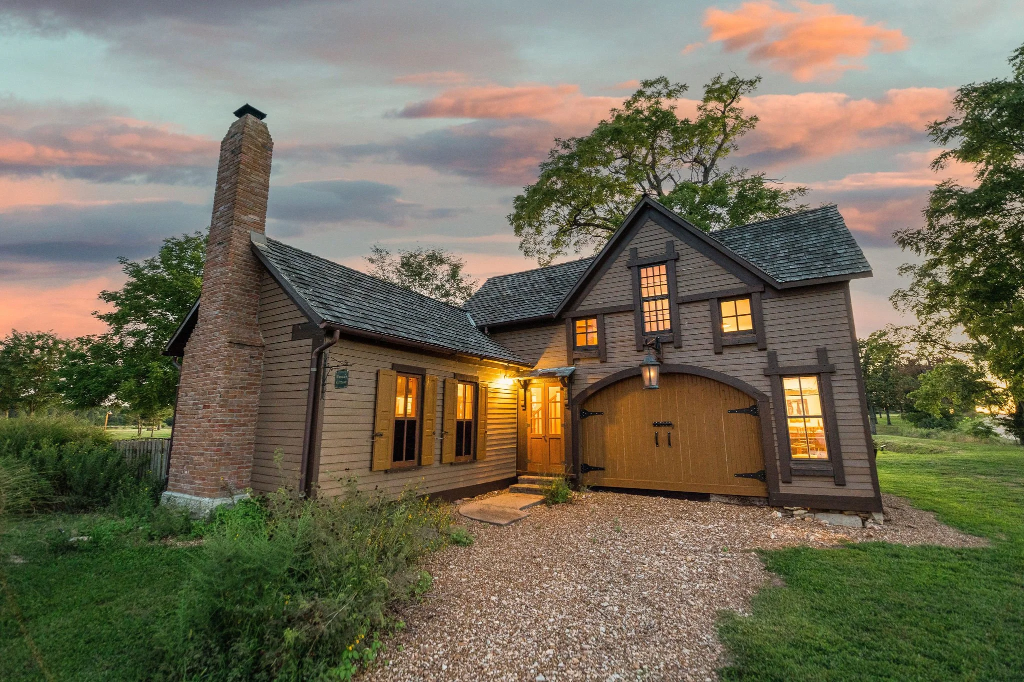 A cozy house with a brick chimney at sunset, with warm interior lights shining through the windows, surrounded by green grass and trees.