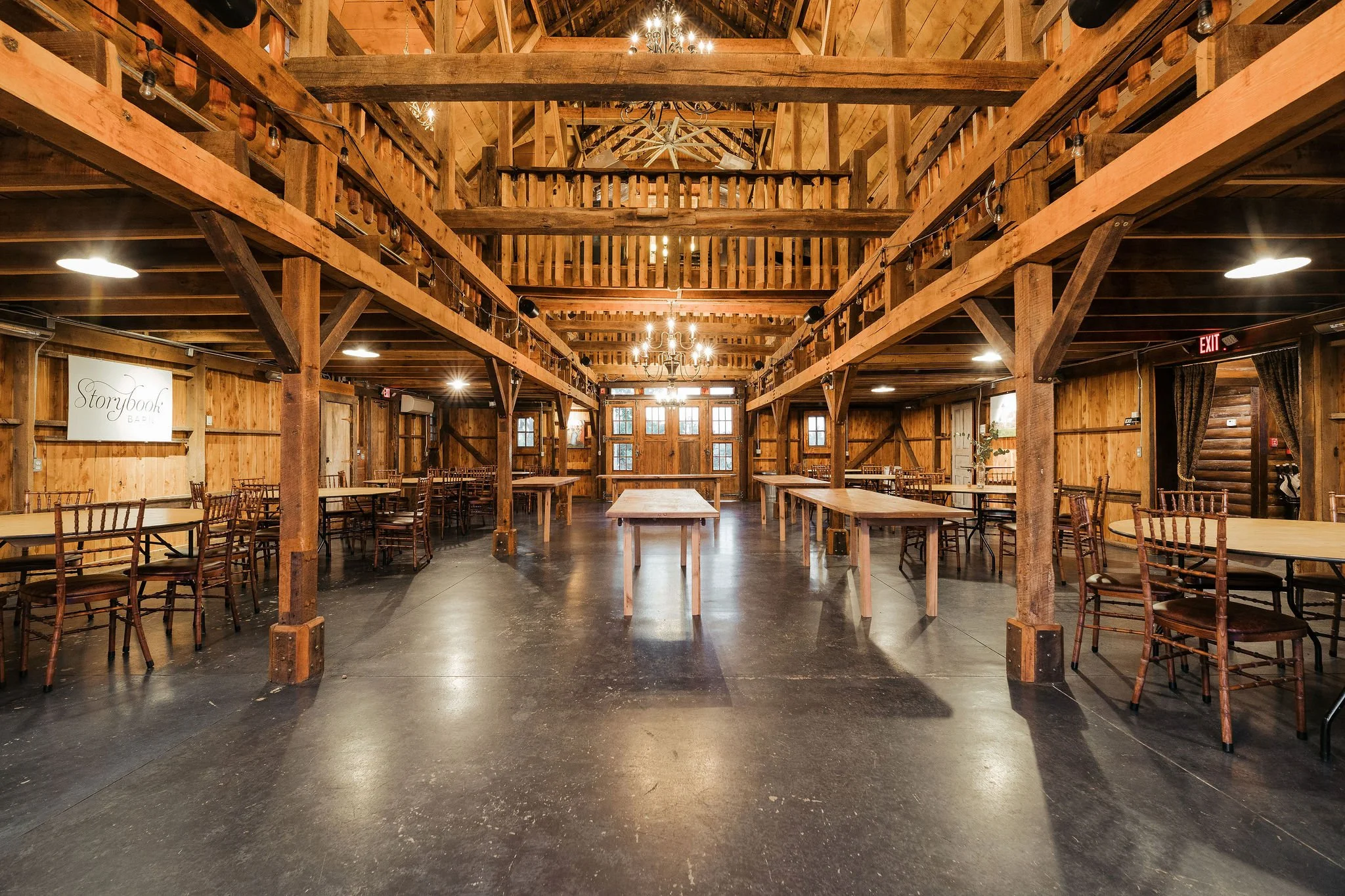 Interior of a rustic event space with wooden walls, ceiling, and beams, featuring multiple tables and chairs, chandeliers, and string lights.