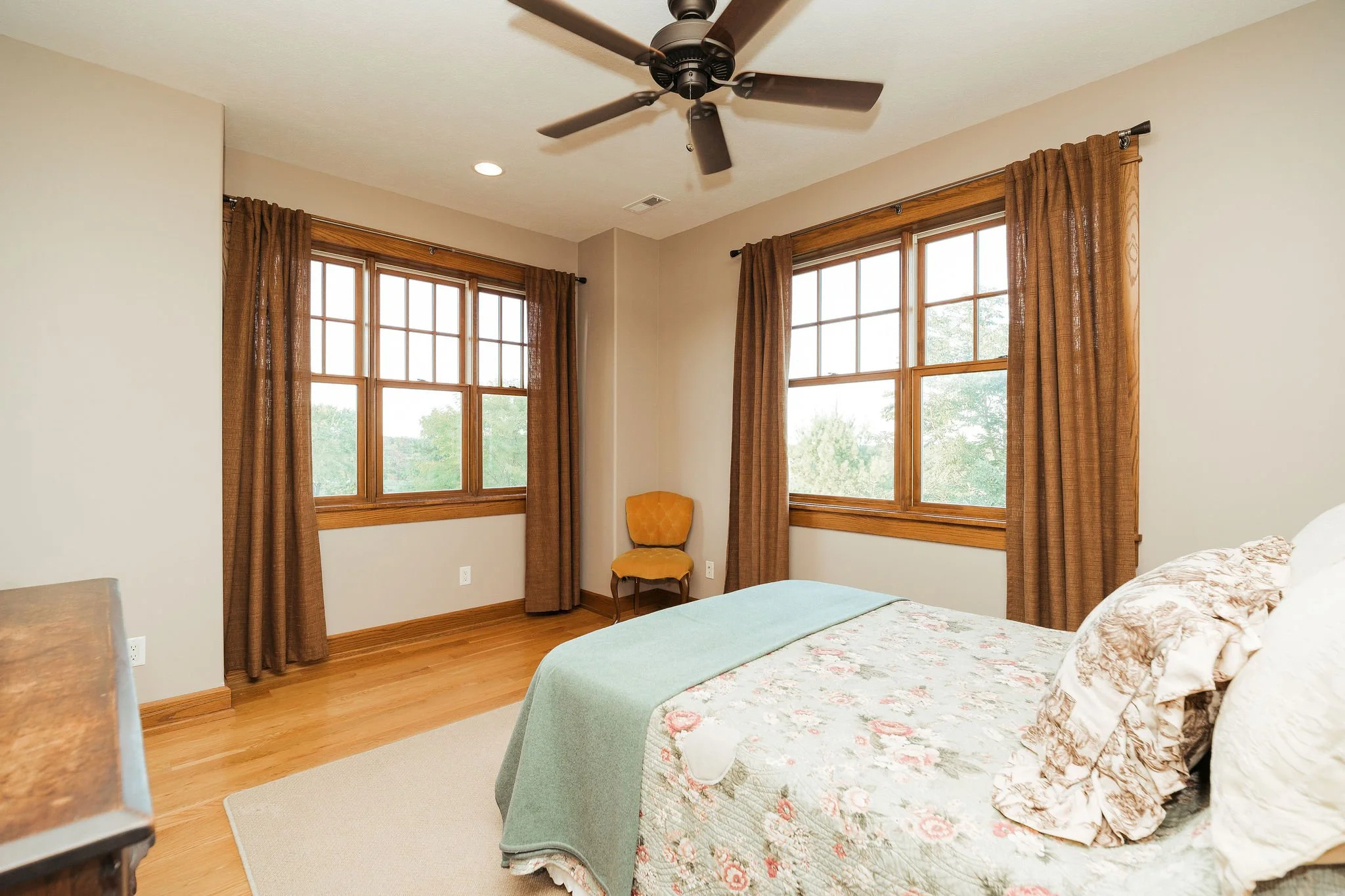 A bedroom with two large windows, brown curtains, a bed with floral bedding, a yellow upholstered chair, and a ceiling fan.