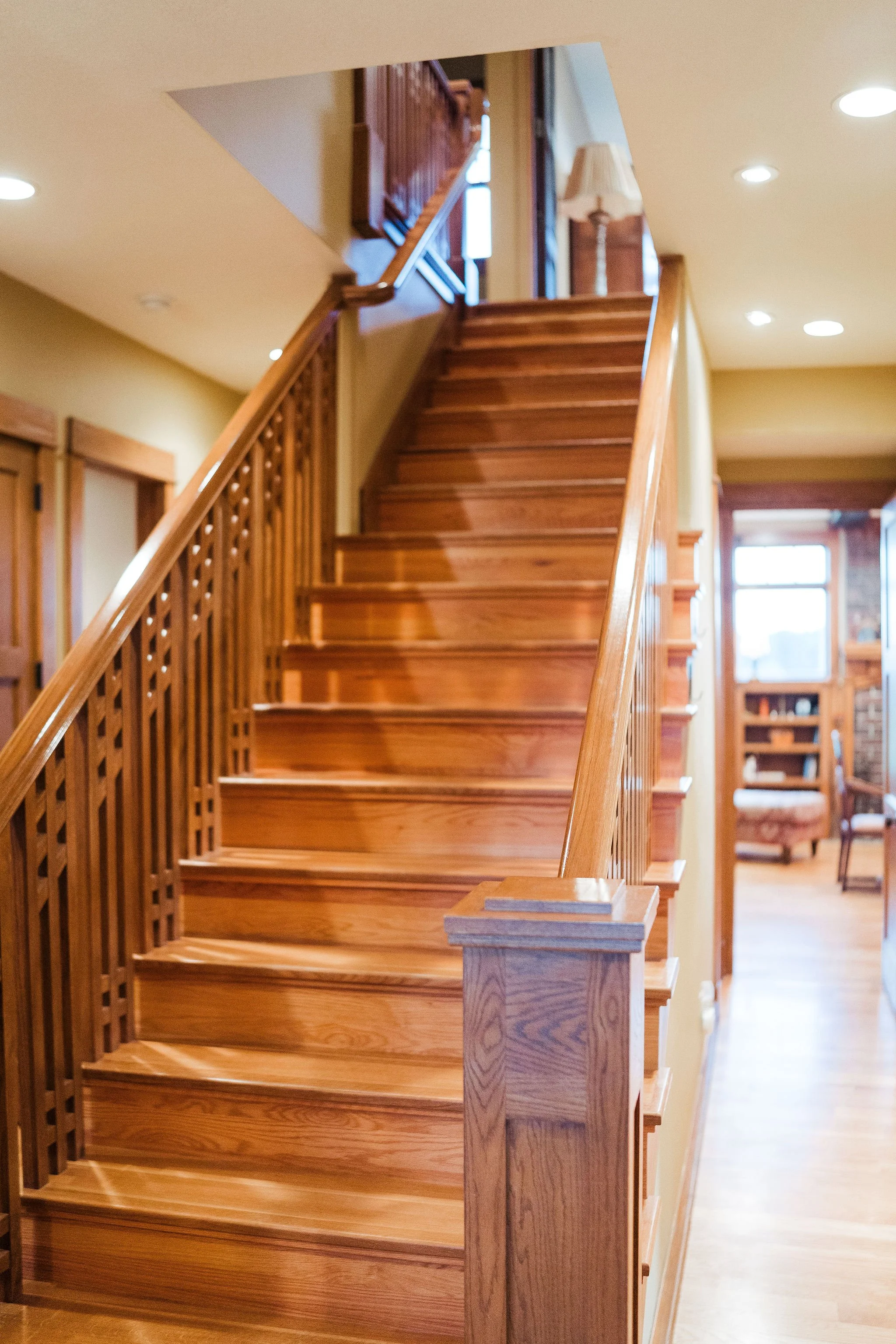 Wooden staircase in a house interior leading to an upper floor with a hallway and furniture in the background.