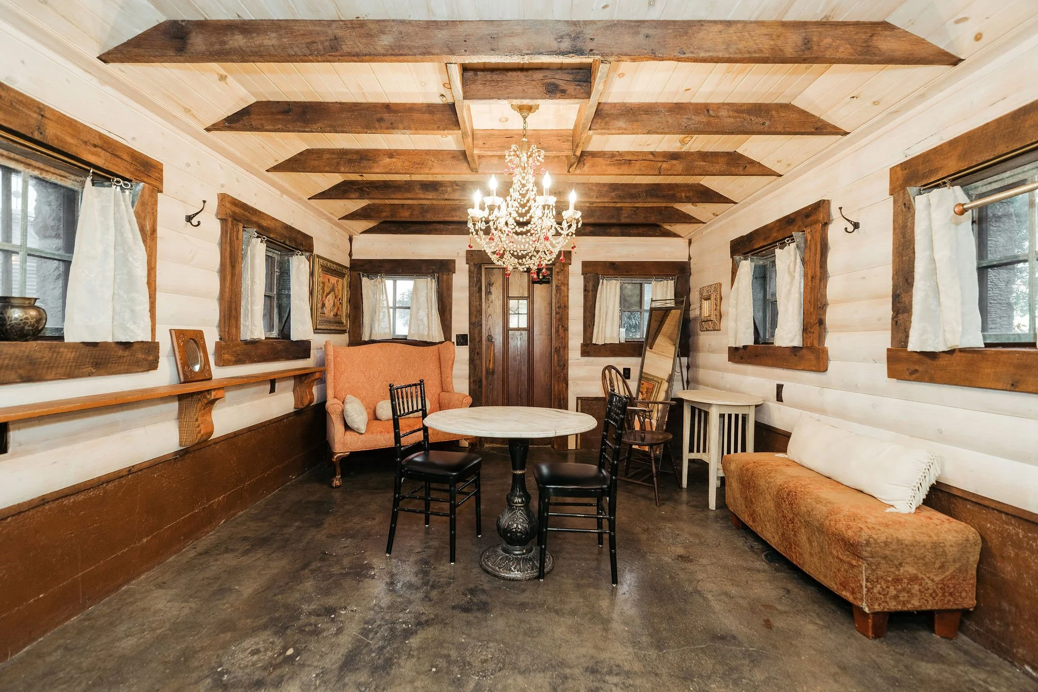 Interior of a rustic room with wooden walls, ceiling beams, a chandelier, and vintage furniture including a sofa, chairs, and tables.