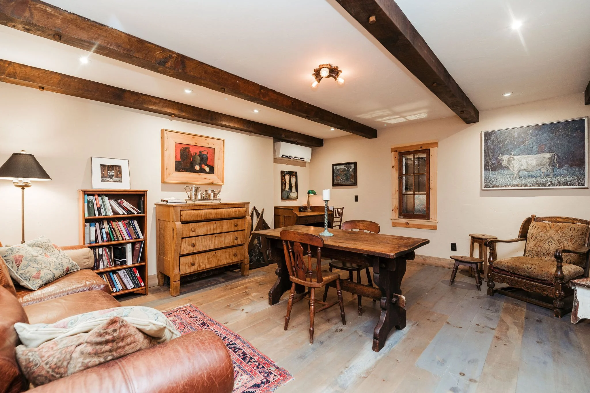 Living room with wooden beams on ceiling, wooden furniture, leather sofa, patterned armchair, bookshelves, and framed paintings on the walls.