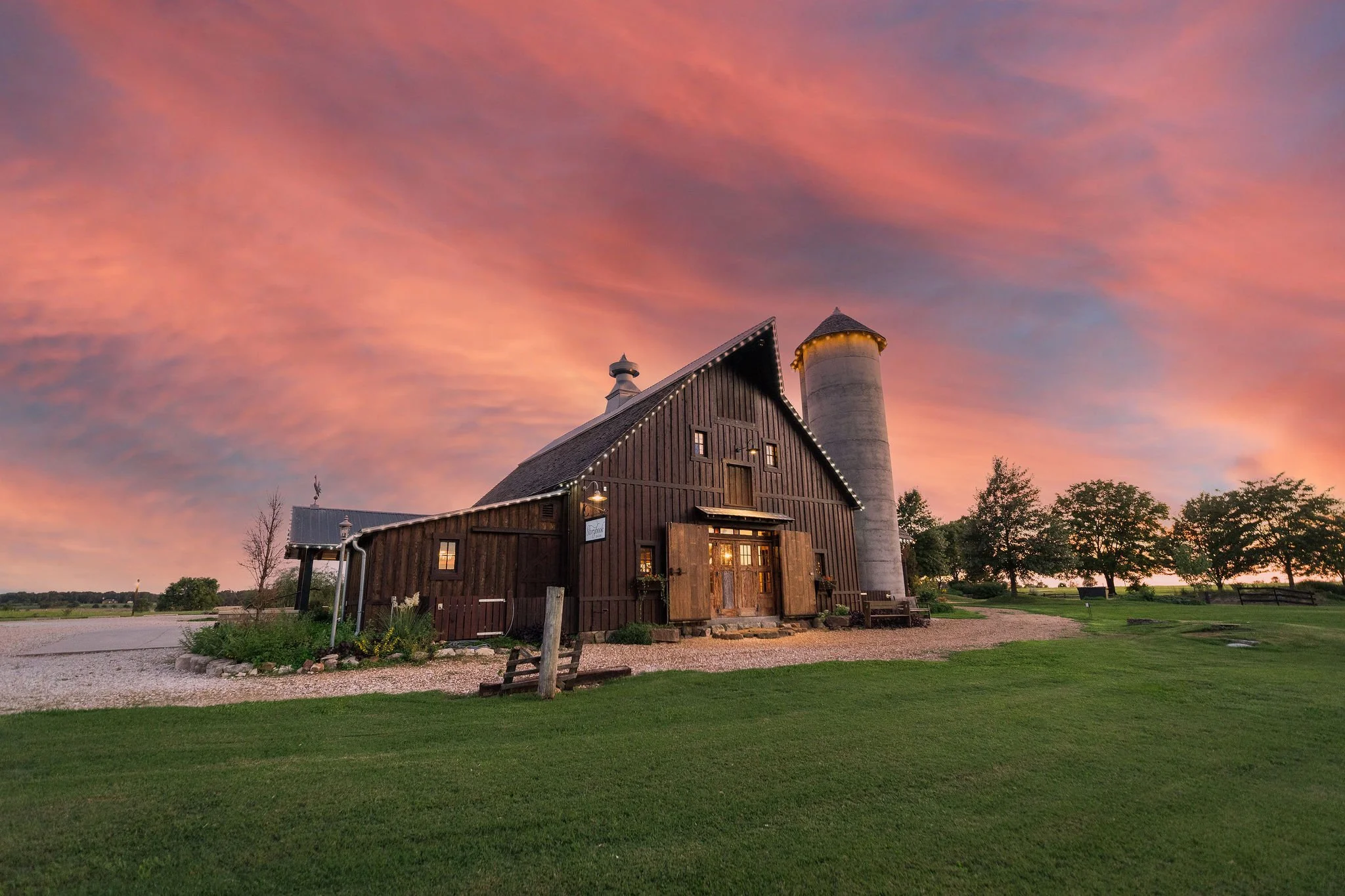 A rustic barn with a silo at sunset, clouds with pink and purple hues, surrounded by green grass and trees.