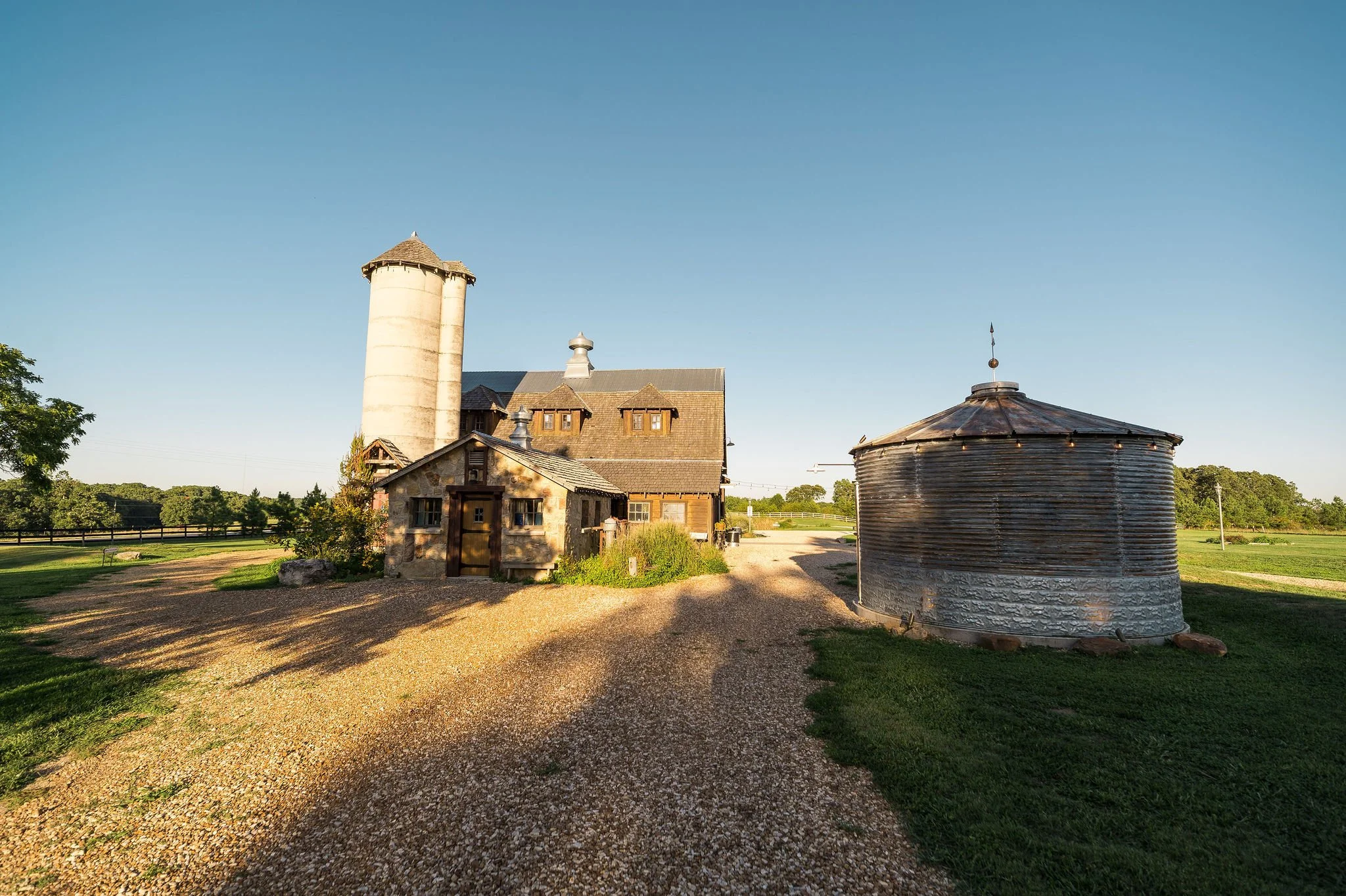A rural farm landscape featuring a stone house with wooden shingles, a tall white silo, and a round wooden water tower under a clear blue sky with green fields in the background.
