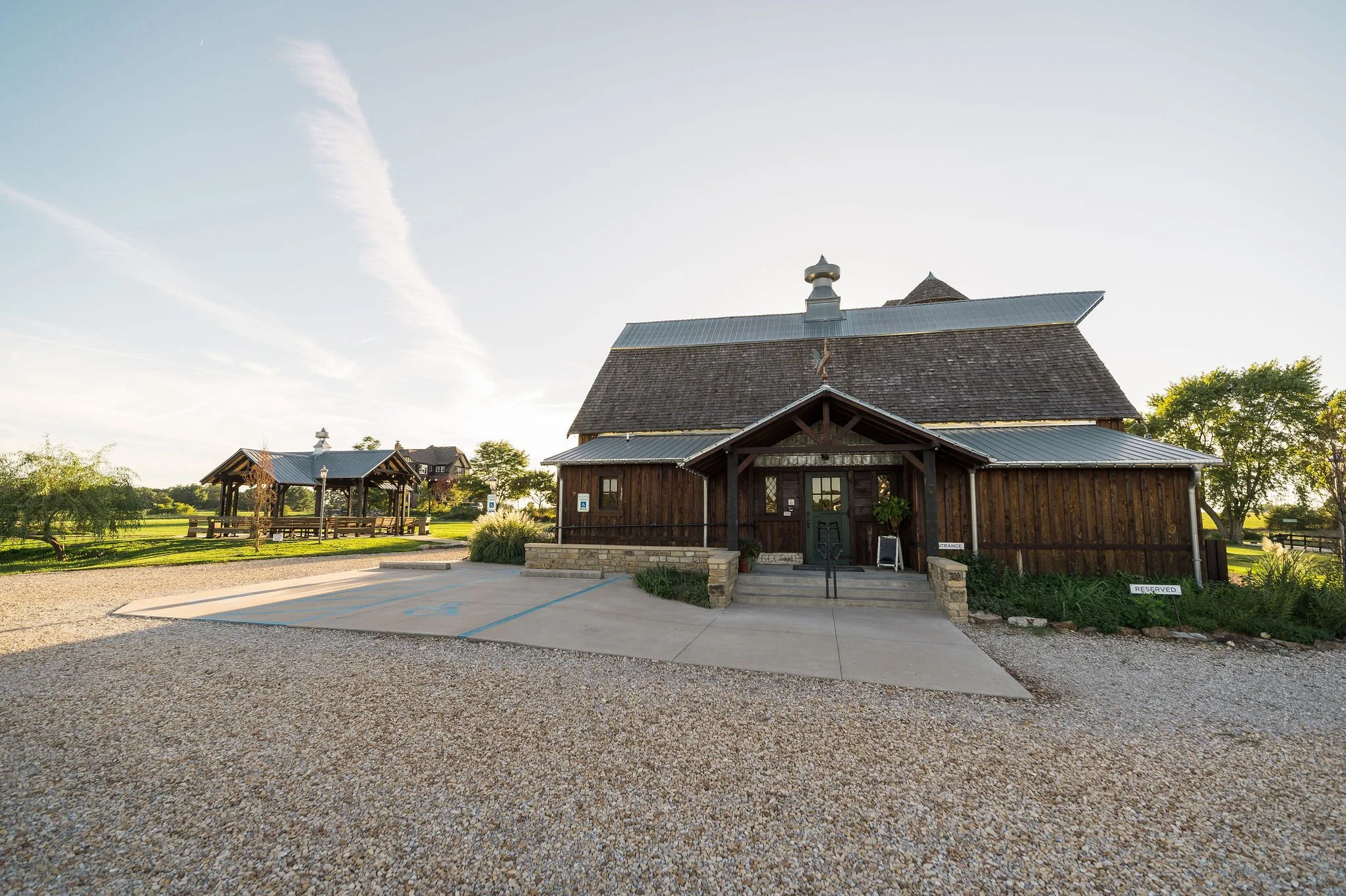 A rustic wooden church with a metal roof, surrounded by greenery and a gravel parking lot, under a clear sky.