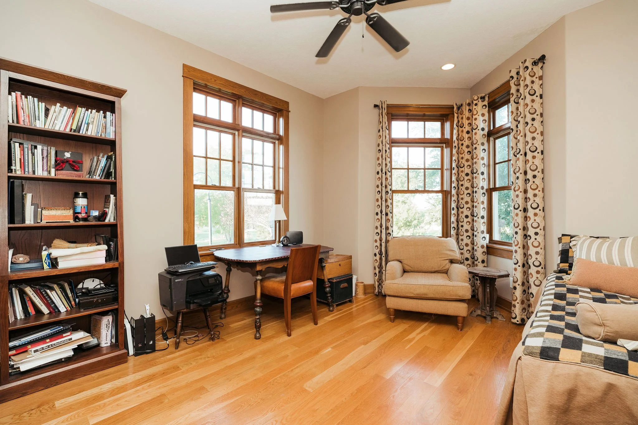 Room with two large windows, a wooden bookshelf filled with books, a small wooden desk with a chair, a beige armchair, a small side table, a patterned curtain, and a ceiling fan.