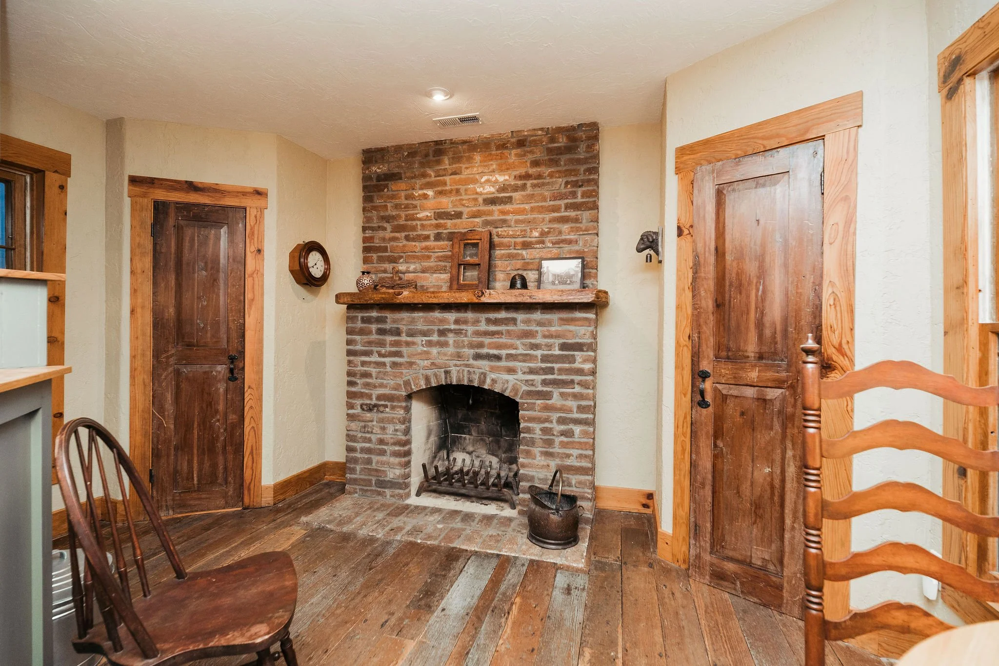 A cozy living room corner with a brick fireplace and wooden accents. There are two wooden doors, a wooden chair, and decorative items on the mantle, including a clock and framed picture.