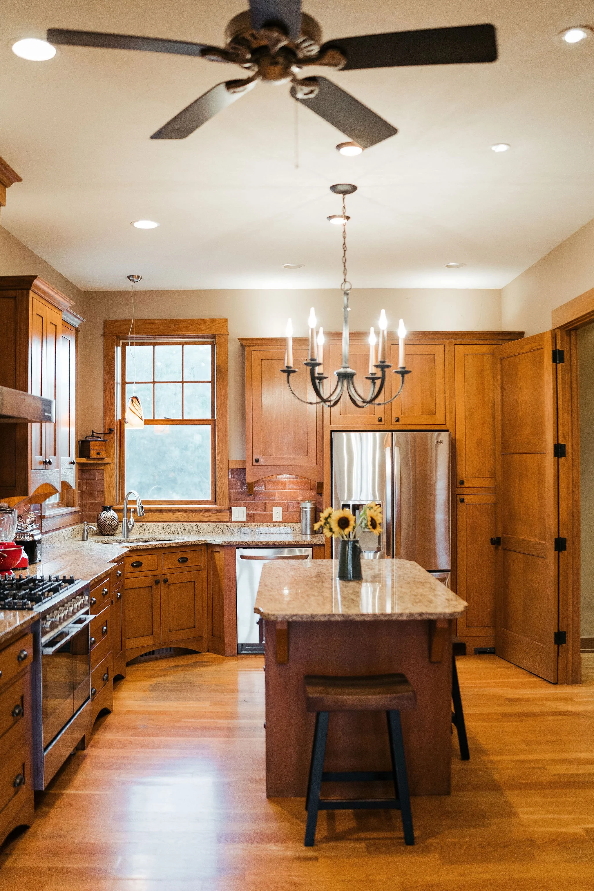 A cozy kitchen with wooden cabinets, granite countertops, stainless steel appliances, hanging chandelier, ceiling fan, wooden flooring, and sunflower vase on an island.
