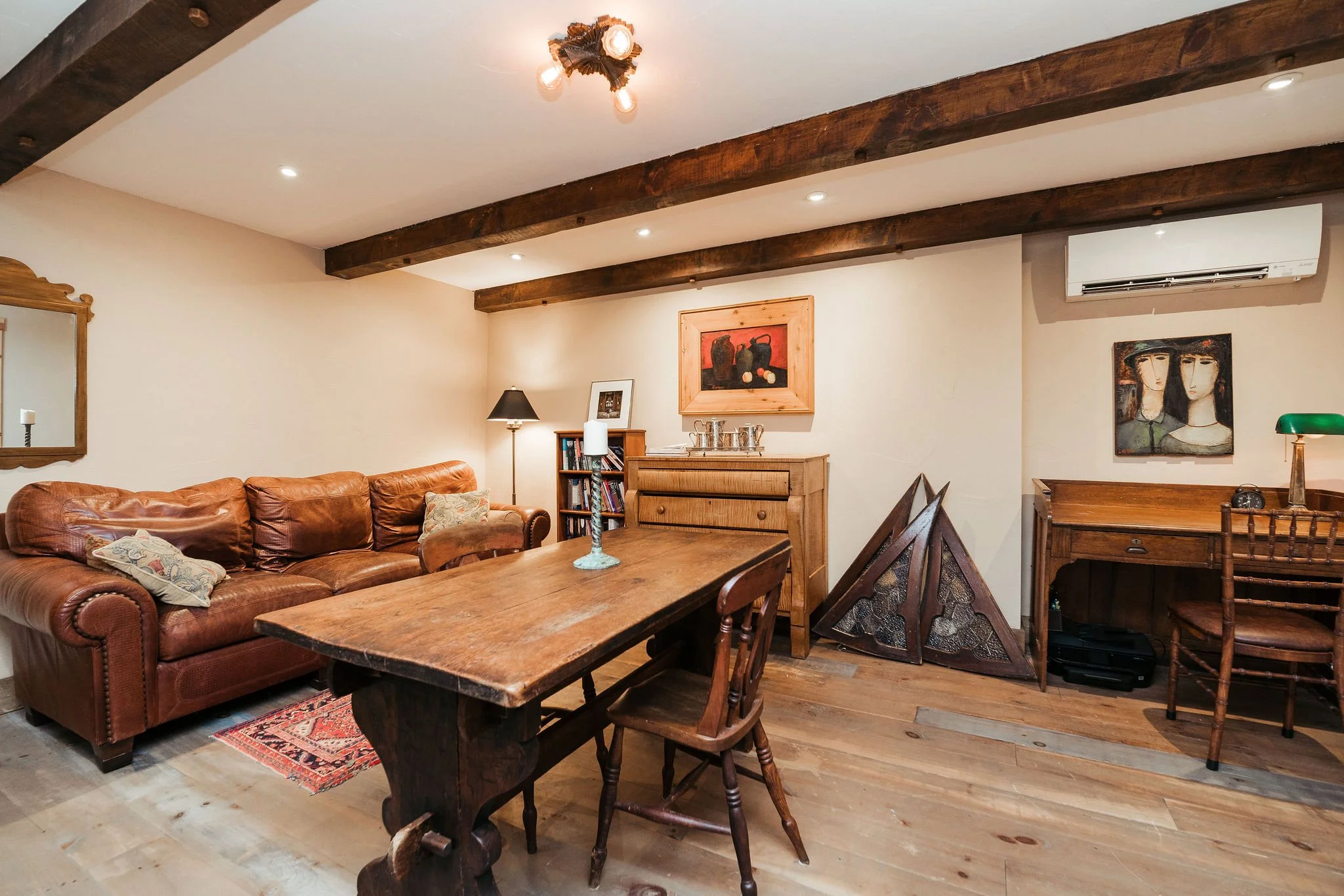 Living room with beige walls, wooden beams on the ceiling, leather sofa, wooden table with a candle holder, desk with a green banker-style lamp, framed artwork on the walls, air conditioning unit, and hardwood floor.