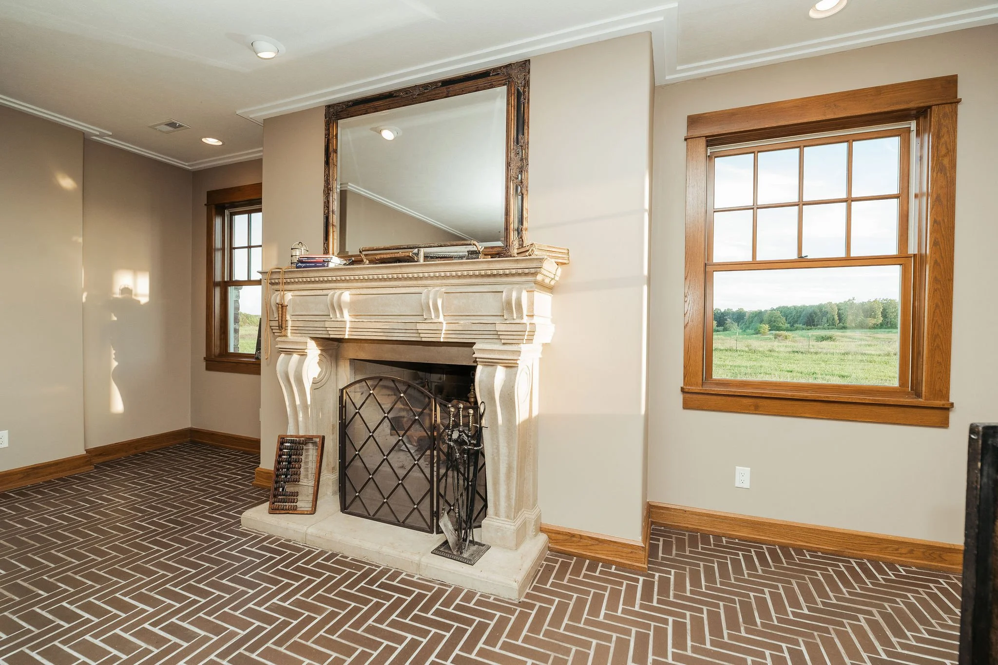 Living room with brick-patterned flooring, a fireplace with a decorative mantel, a large wooden framed mirror above the mantel, and two large windows showing a green landscape outside.