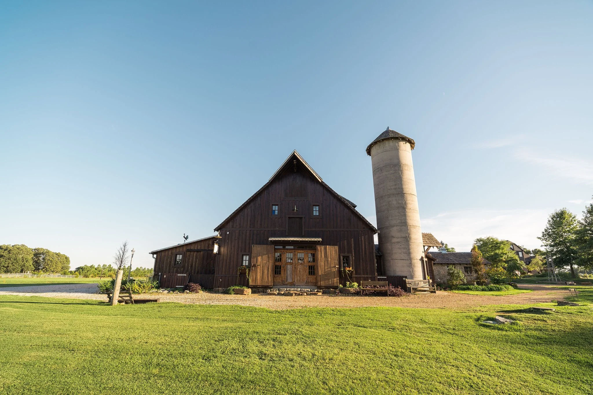 A rustic barn with a tall silo on a farm, surrounded by green grass and trees under a clear blue sky.
