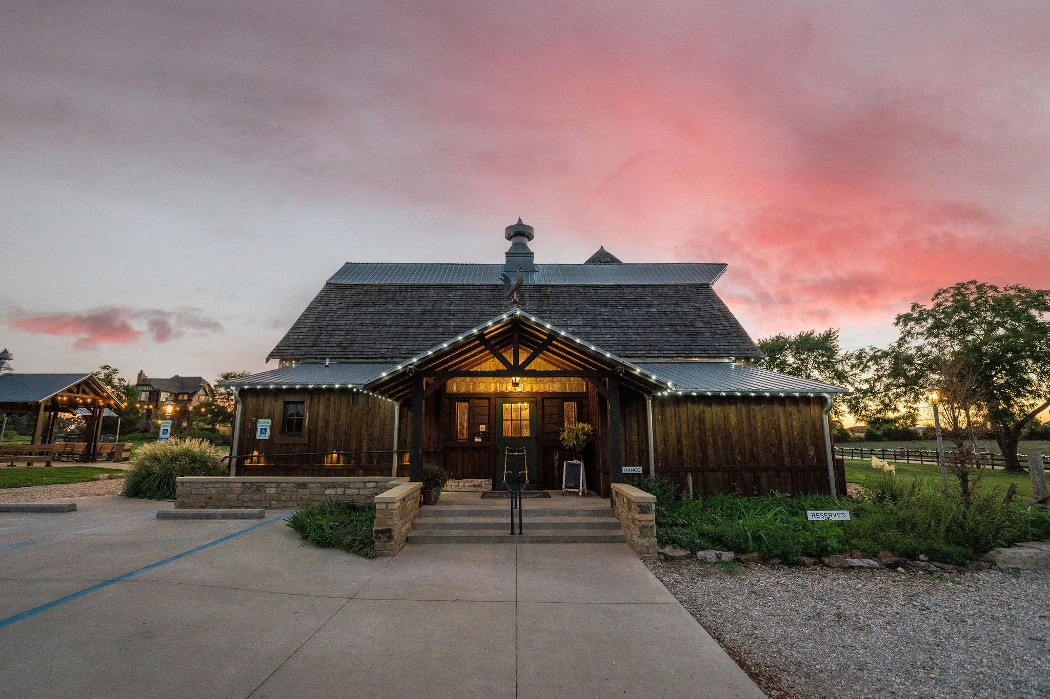 A rustic wooden building decorated with string lights at sunset, with a concrete walkway leading to steps at its front entrance, landscaped bushes, and signs indicating reserved and accessible parking.