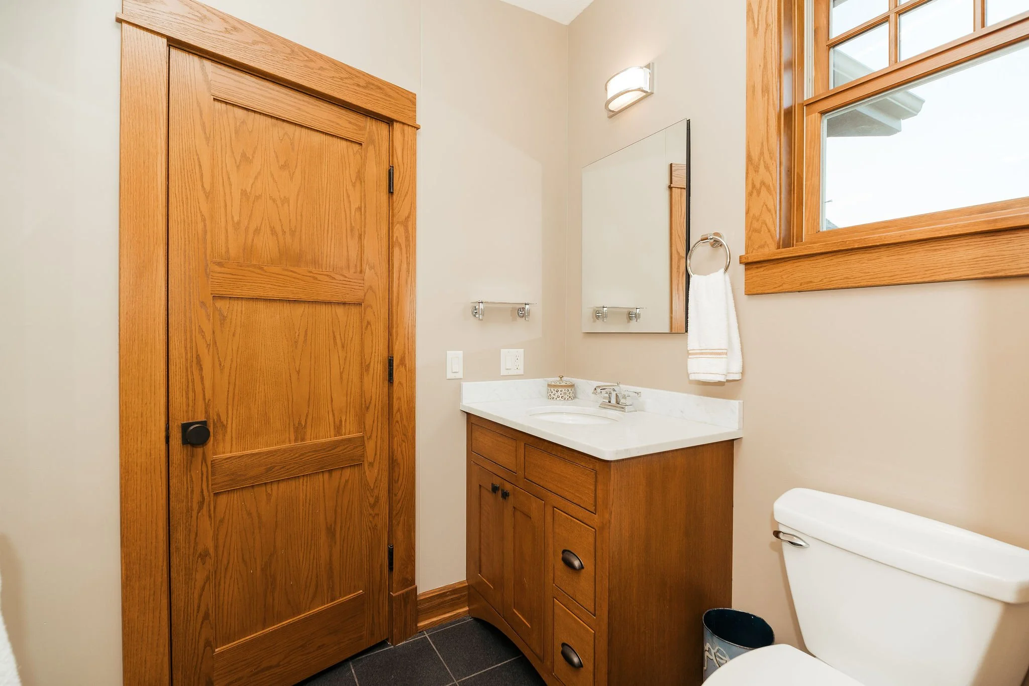 Bathroom with wooden door, wooden vanity with white marble countertop, mirror, white towel, and window with wooden trim.