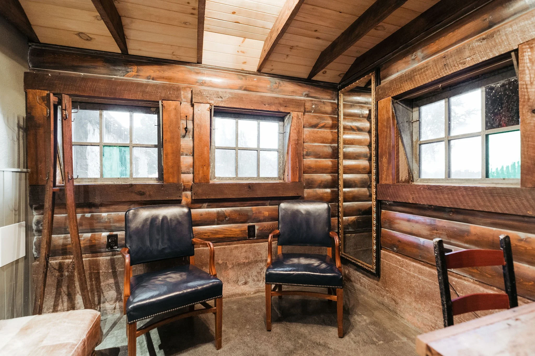 Interior of a rustic room with wooden walls and ceiling, two vintage black leather chairs, and two small windows with wooden frames.