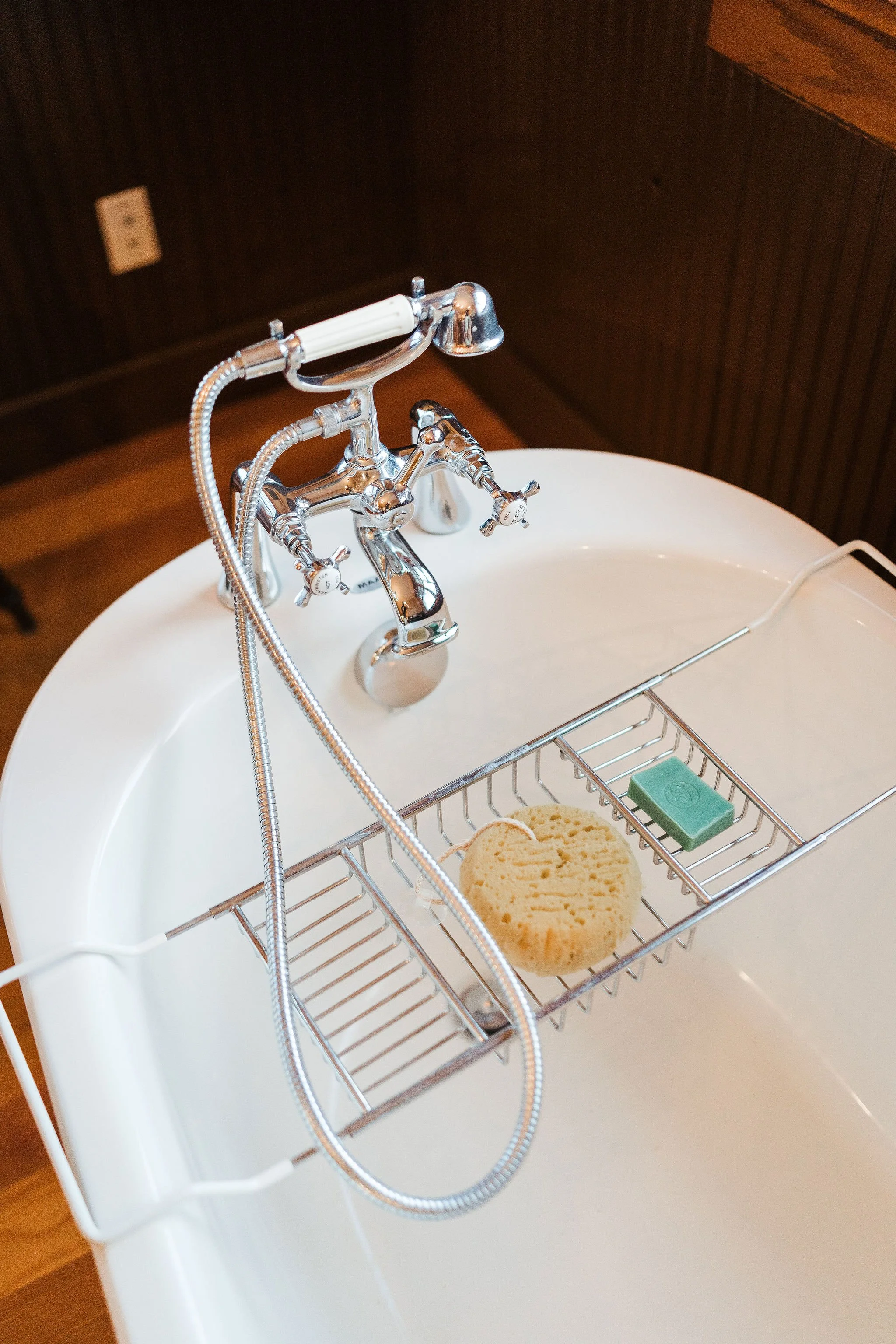 Over-the-bathtub metal rack with a sponge, a green soap bar, and a shower hose attached to a chrome faucet in a bathtub.