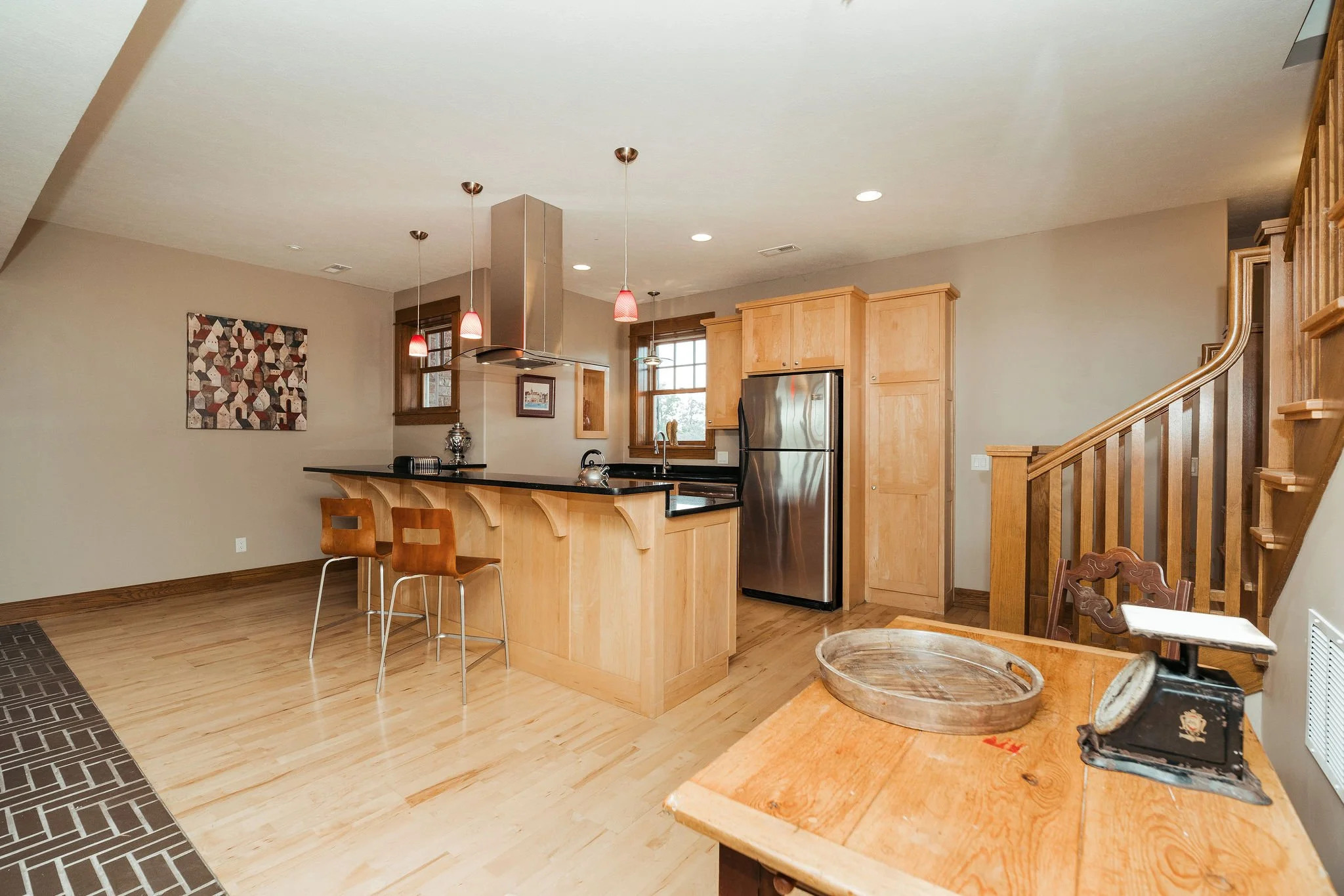 Kitchen with wooden cabinets, island with black countertop, stainless steel refrigerator, wooden staircase, and vintage scale on a wooden table.