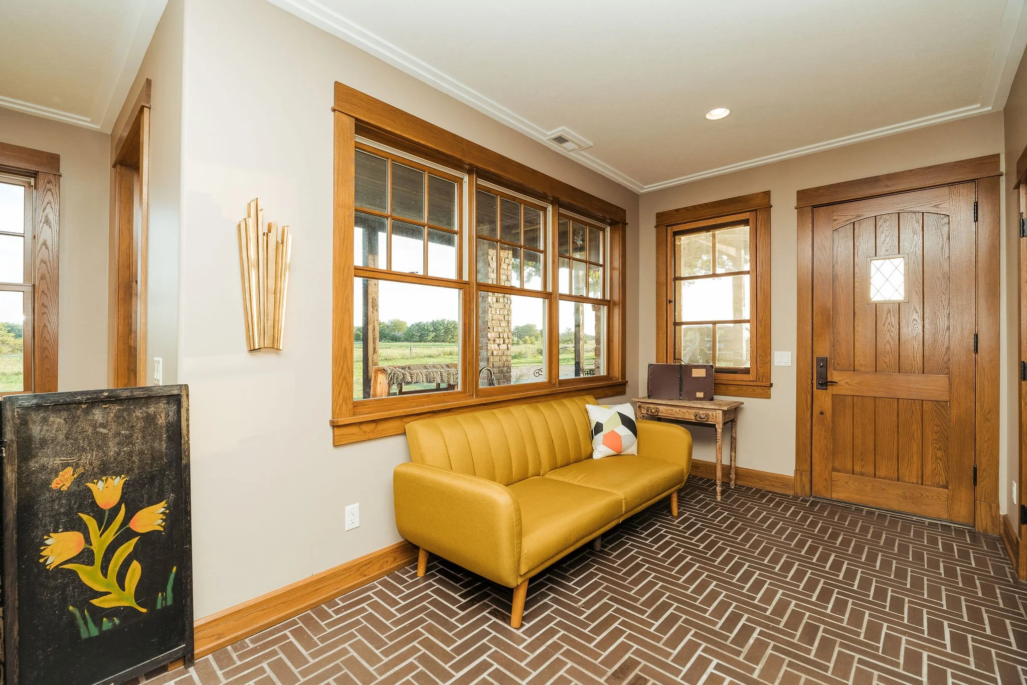 Living room with large windows, wooden trim, mustard-colored sofa, decorative pillow, wooden door, and artwork, with a view of green landscape outside.
