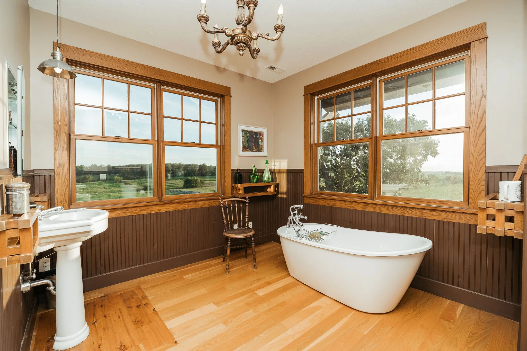A spacious bathroom features a freestanding white bathtub, a vintage wooden chair, large windows with wooden trim, and a small wall shelf with green glass bottles. The walls are partially paneled with dark wood wainscoting and the floor is light hard