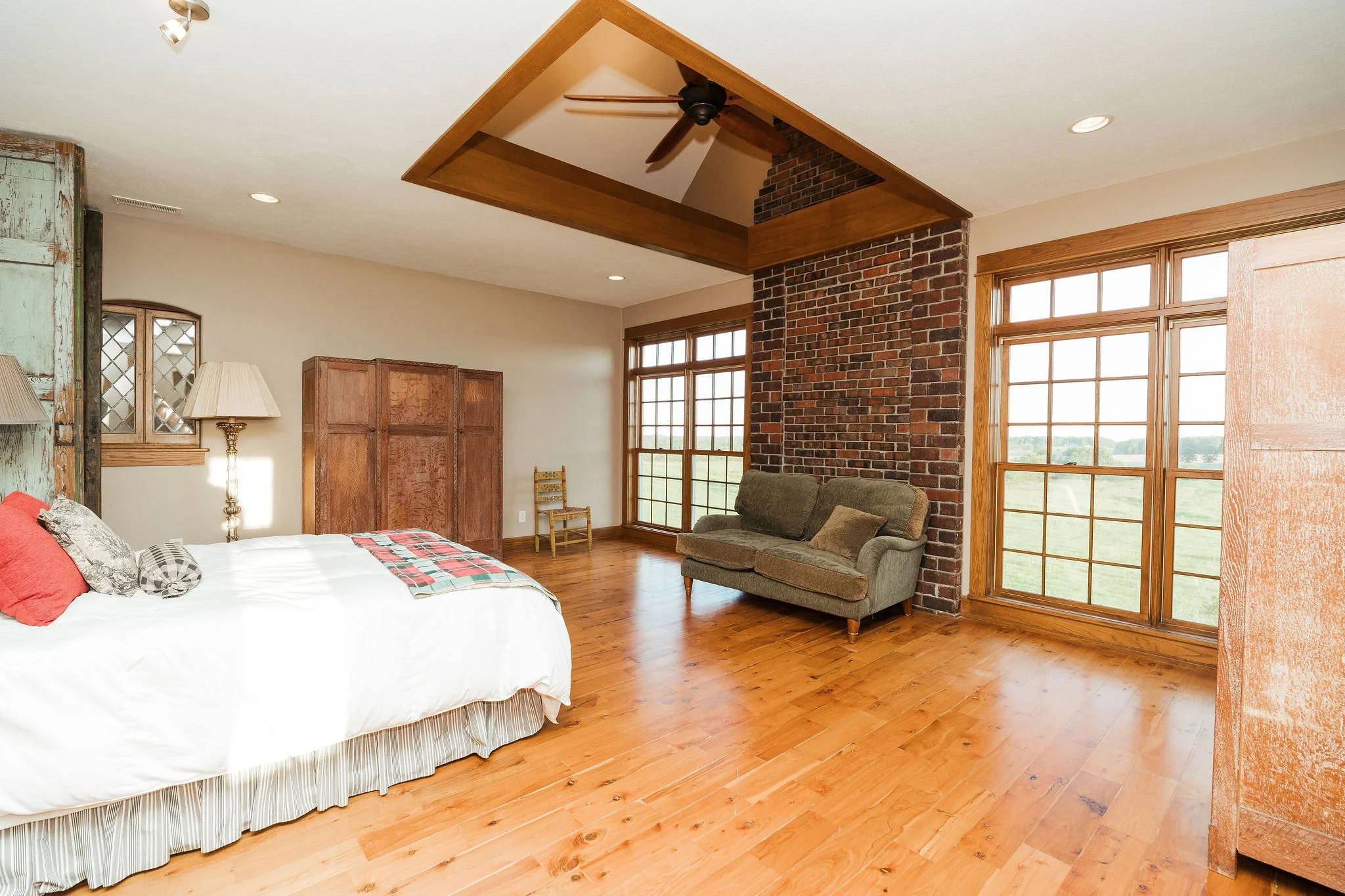 A spacious bedroom with large windows, a brick chimney, and wooden flooring. The room includes a bed with pillows, a green sofa, a wooden wardrobe, and a lamp.