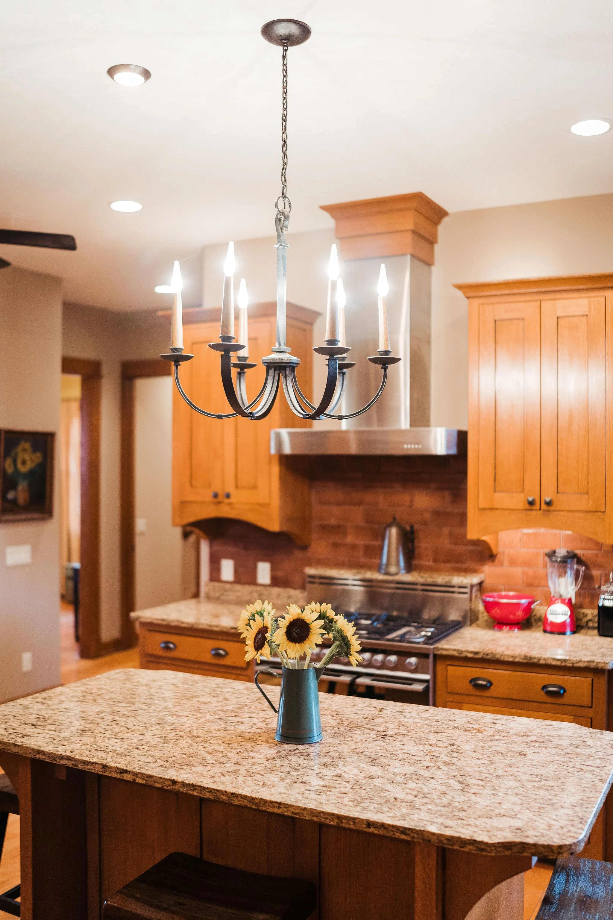 A kitchen with wooden cabinets, a granite countertop, a gas stove with a stainless steel range hood, a vase with sunflowers on the island, and a chandelier hanging from the ceiling.