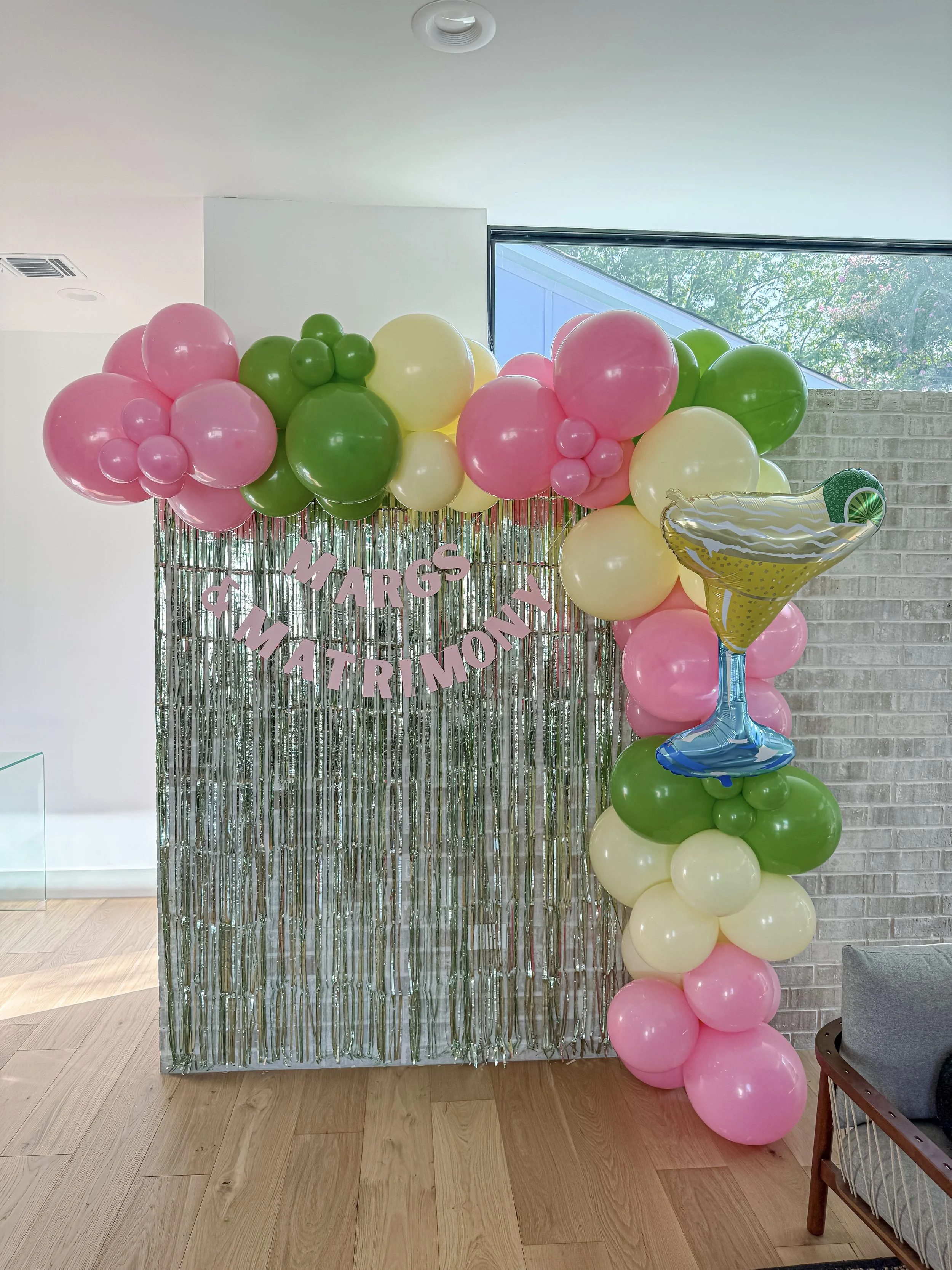 Decorative balloon backdrop with pink, green, and cream balloons, silver fringe curtain, and a pink banner that says "MARGS & MARIAM MARRIAGE," featuring a large inflatable champagne glass balloon.