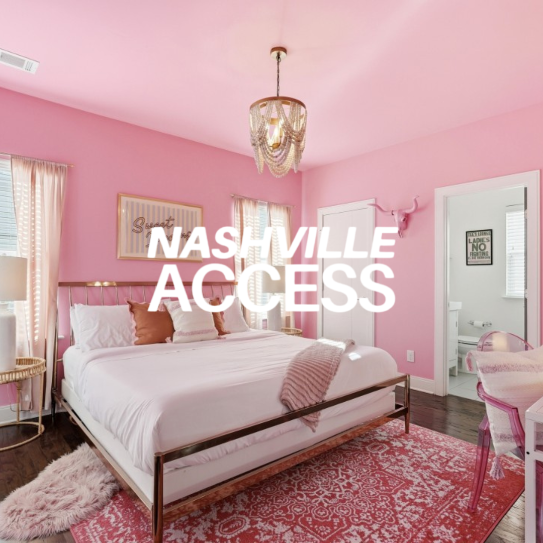 A bedroom with pink walls, white bedding, a pink and white area rug, and pink curtains. There is a chandelier hanging from the ceiling, a pink decorative deer head on the wall, and a small bathroom visible through an open door.
