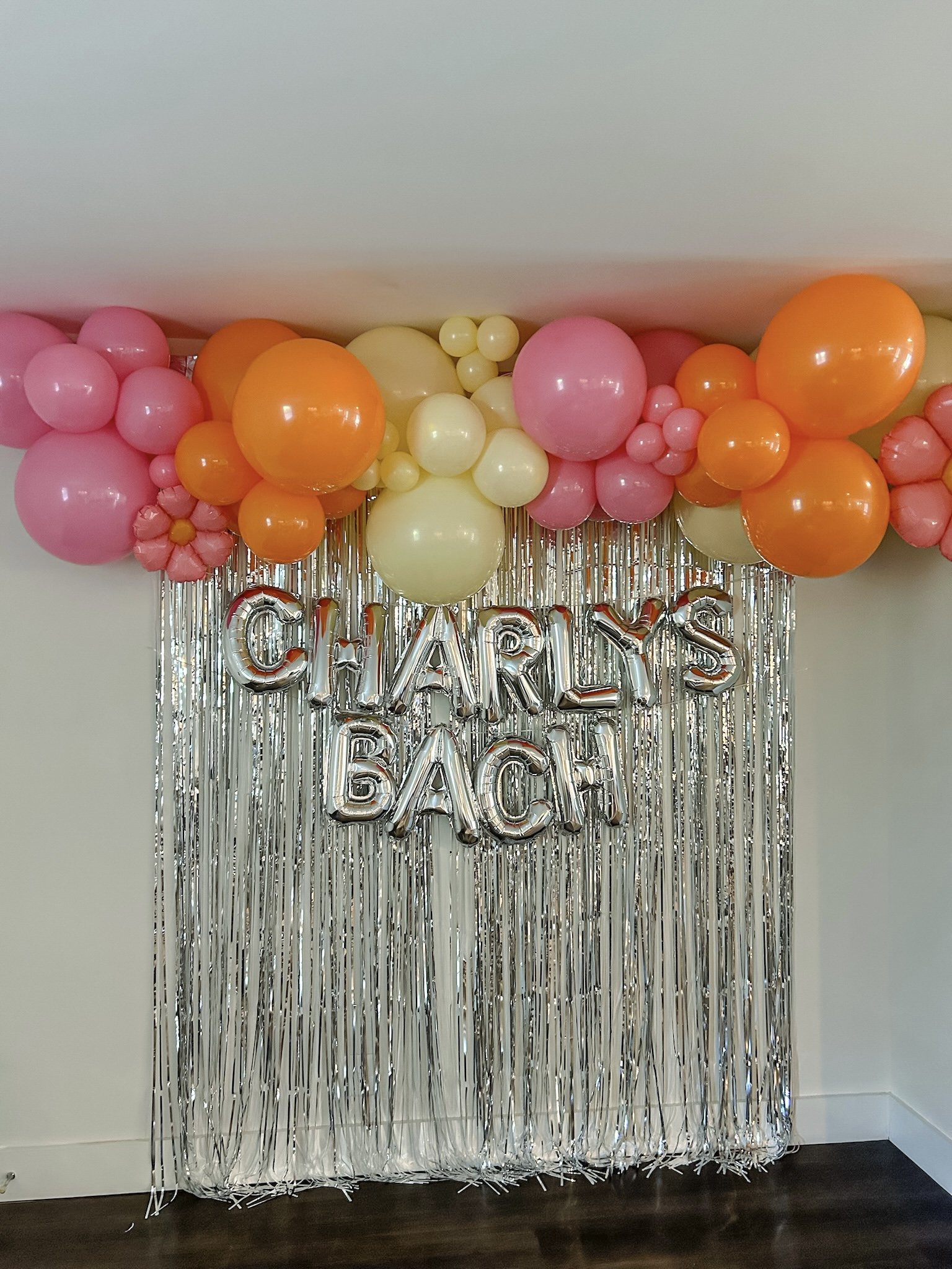 Decorative backdrop with pink, orange, and cream balloons and metallic silver fringe, with silver balloon letters spelling 'CHARLY'S BEACH'.