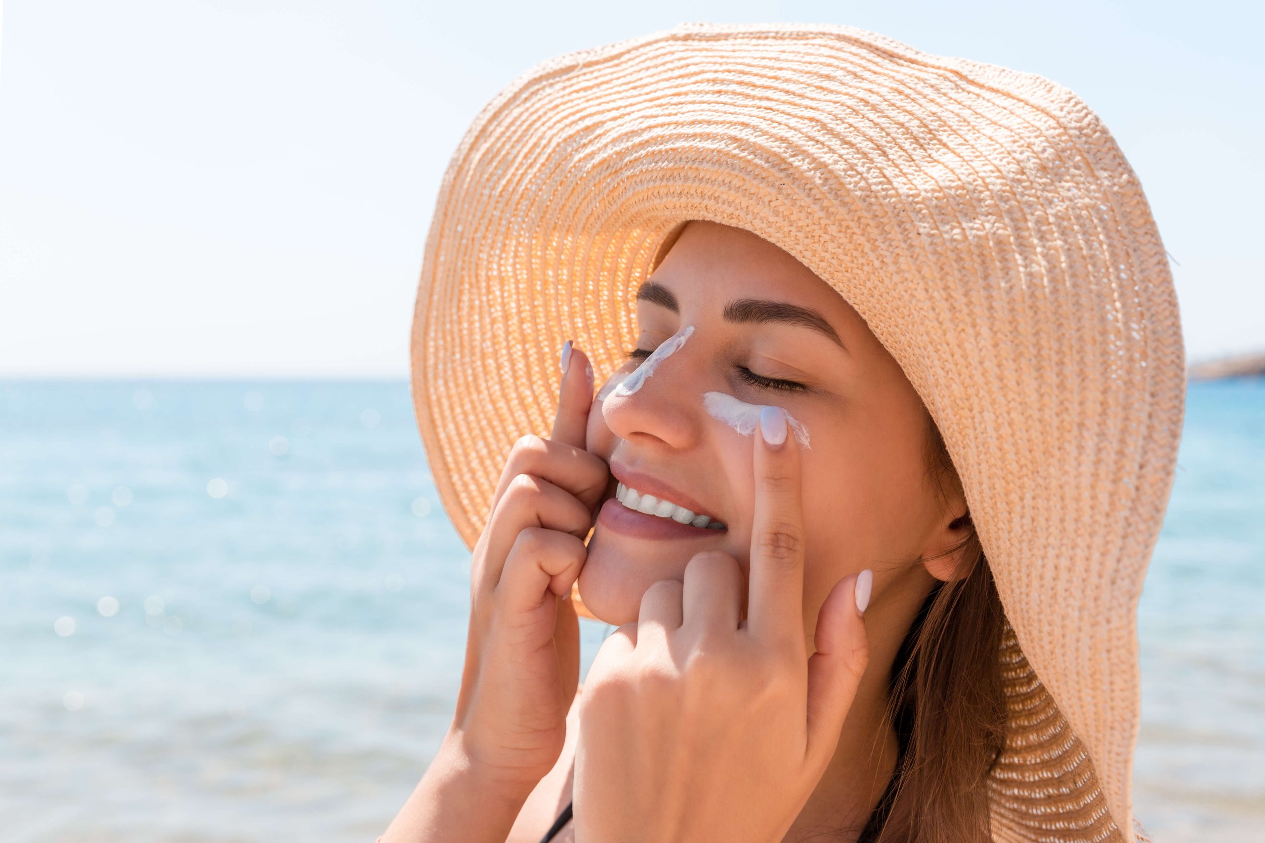 Woman at beach applying sunscreen cream to face while wearing a straw hat.