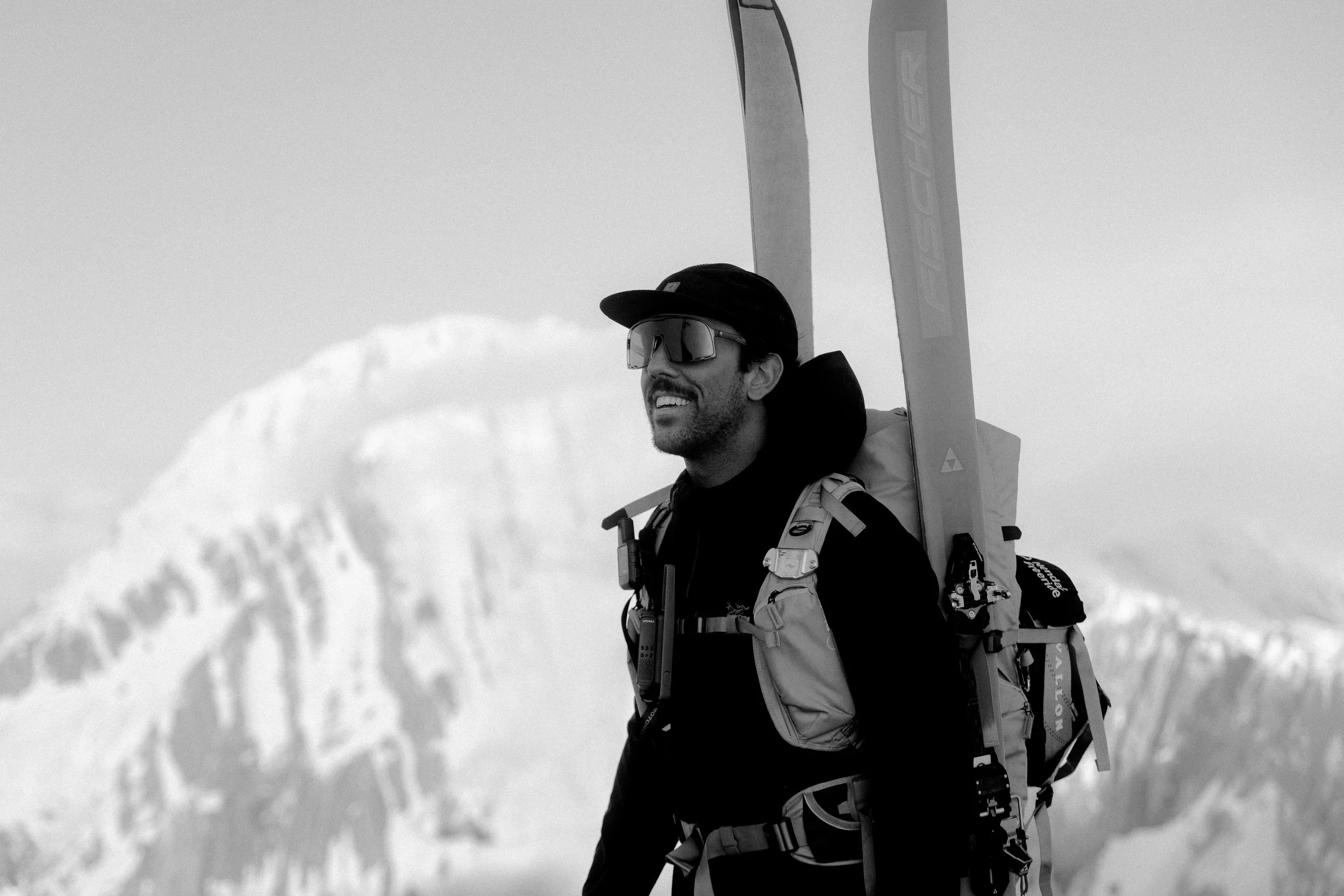 Persona con gafas y gorra lleva esquís en la espalda en un paisaje de montaña nevada.