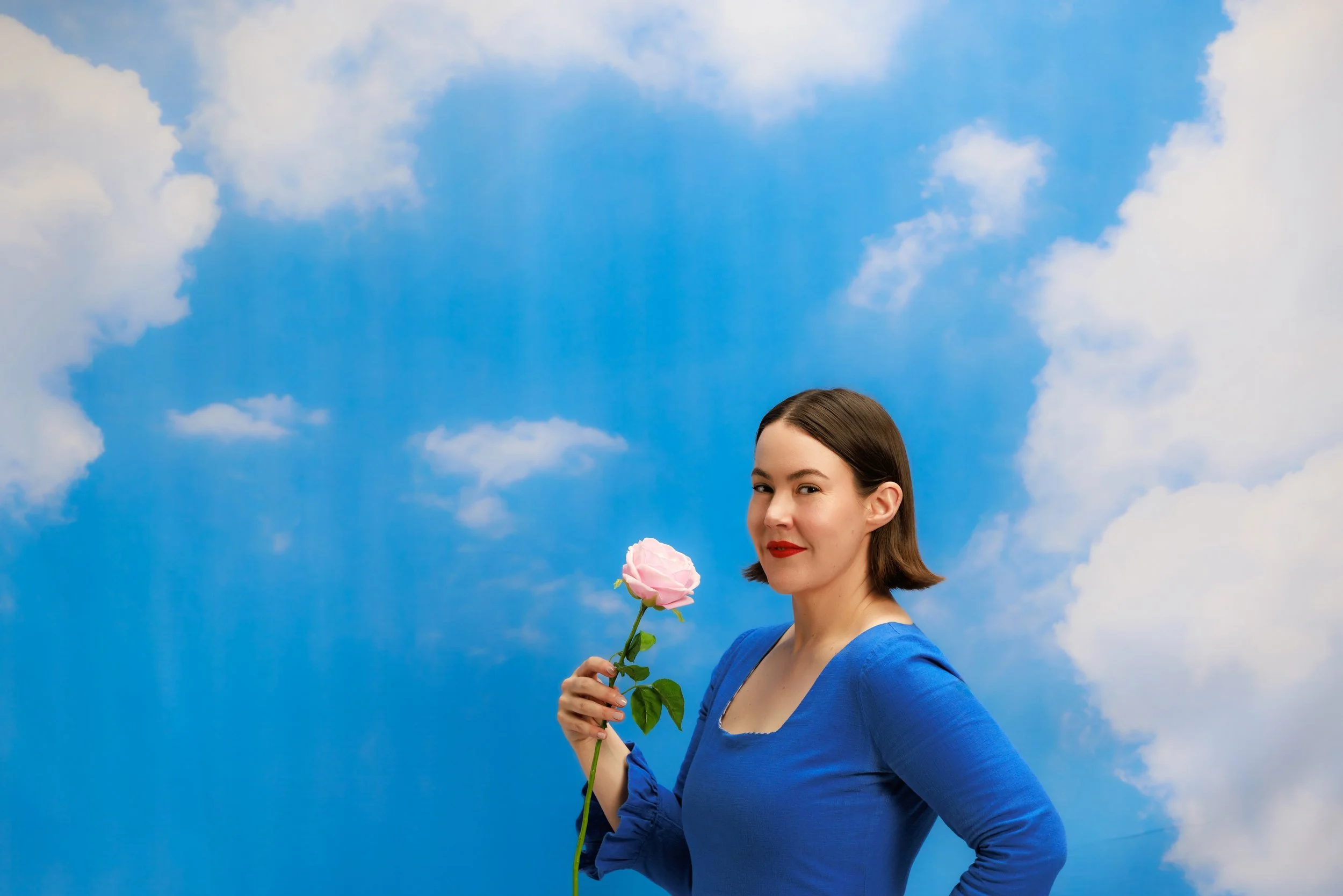 Madeleine Dore in a blue dress holding a pink rose in front of a cloud backgdrop