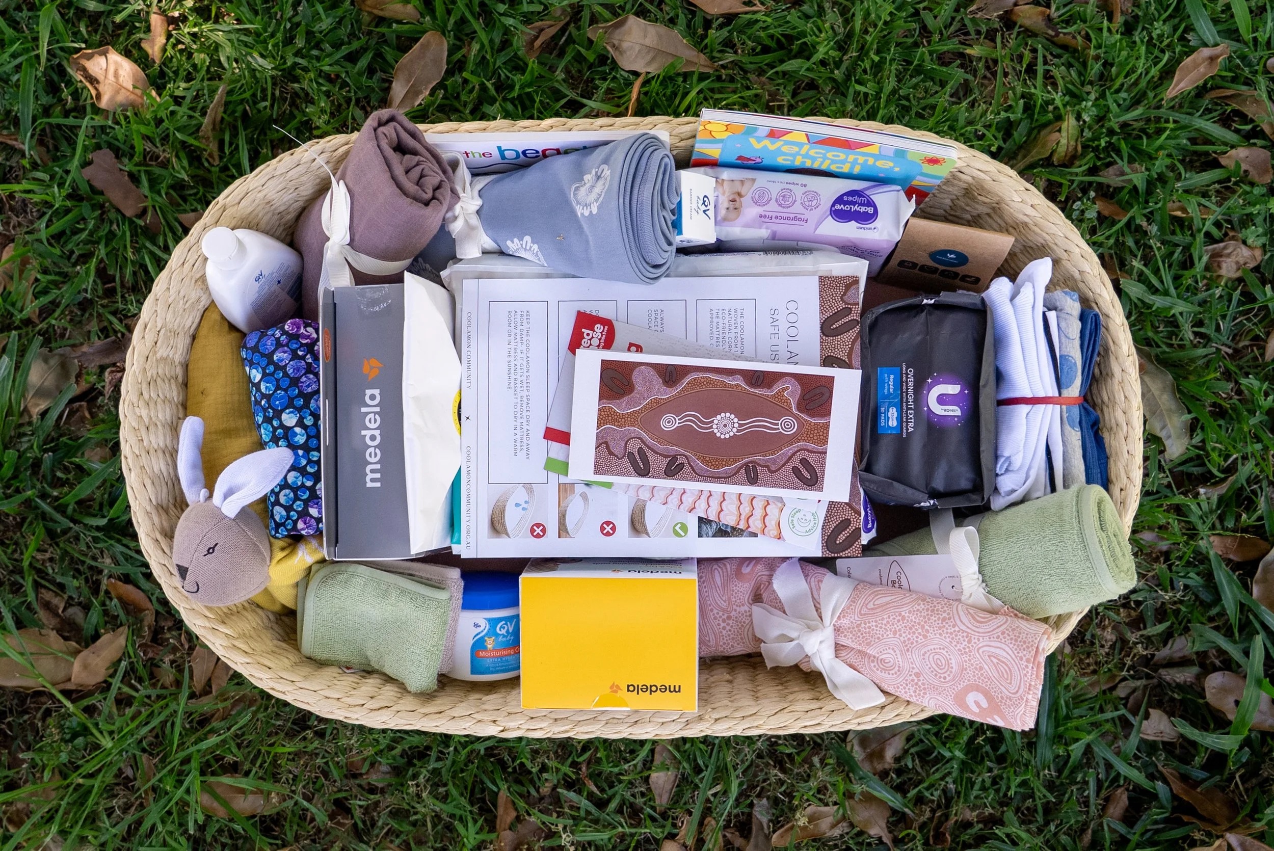 A large woven basket filled with baby clothes, toys, and essentials, with some items spilling out onto a beige surface, against a plain beige background.