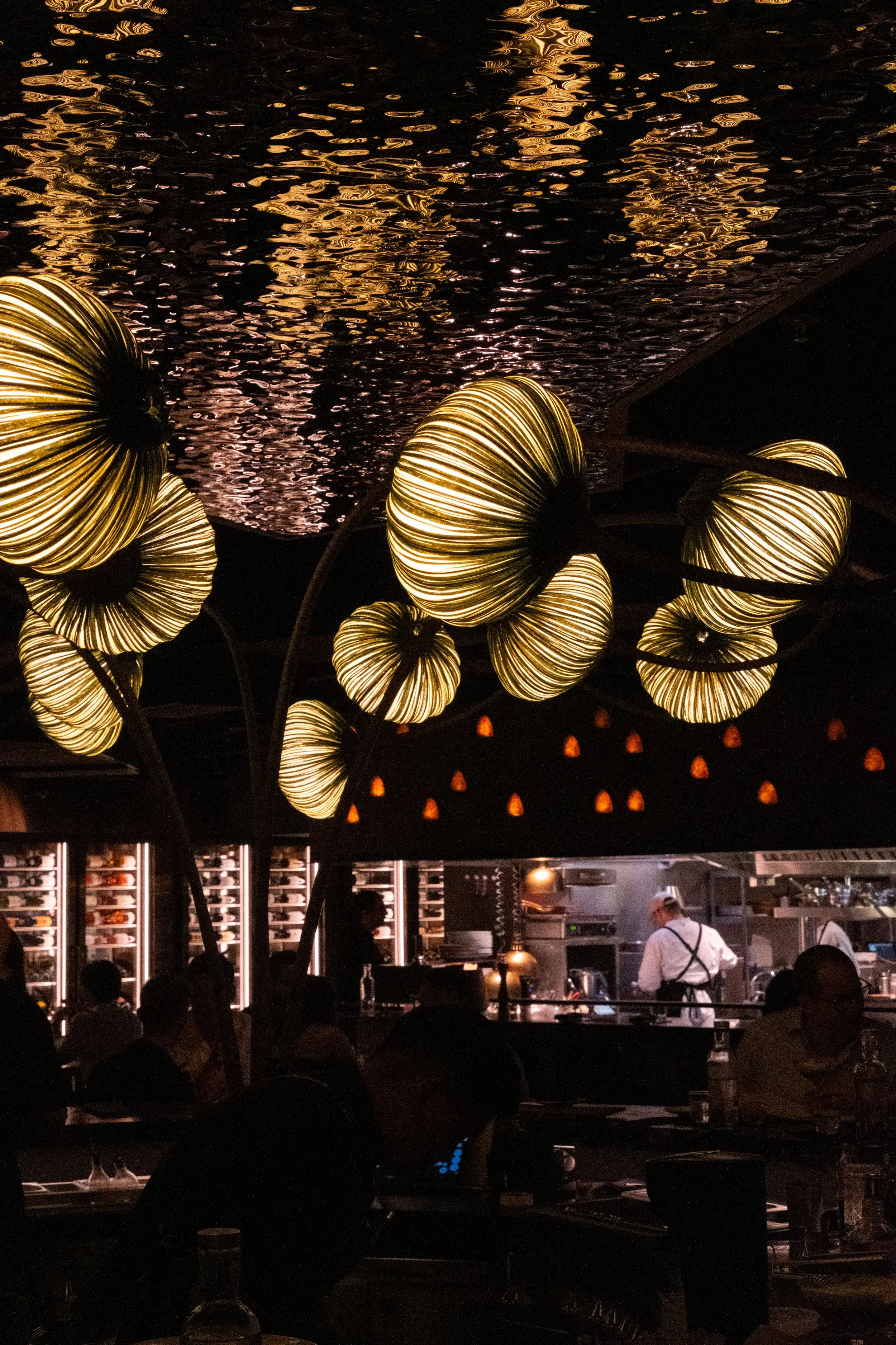 Interior of a restaurant with illuminated decorative lamps resembling large flowers, a reflective water ceiling, and a visible open kitchen in the background.