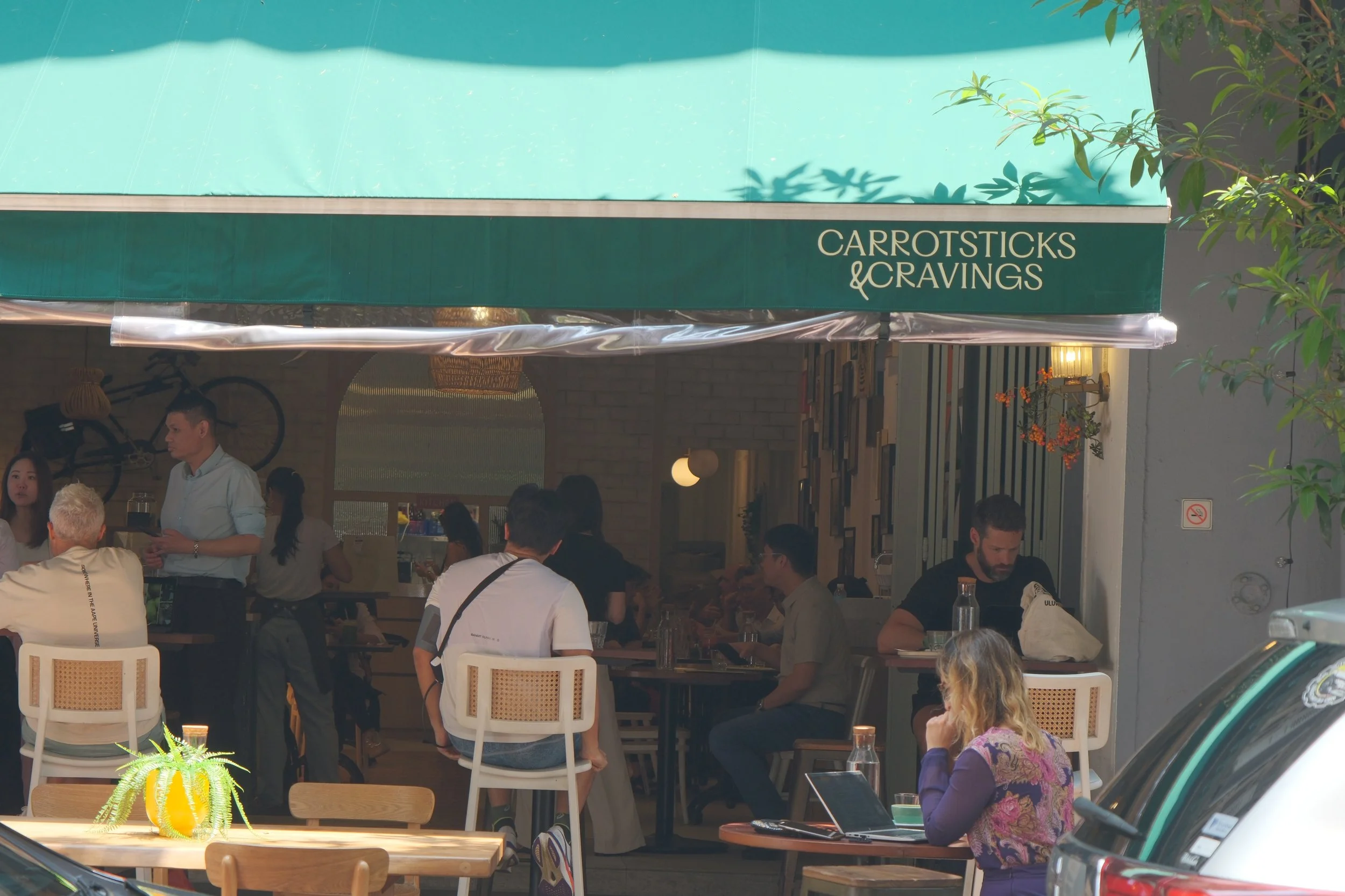 Exterior view of a cafe named 'Carrotsticks & Cravings' with people sitting and standing near the entrance, some working on laptops and others talking.
