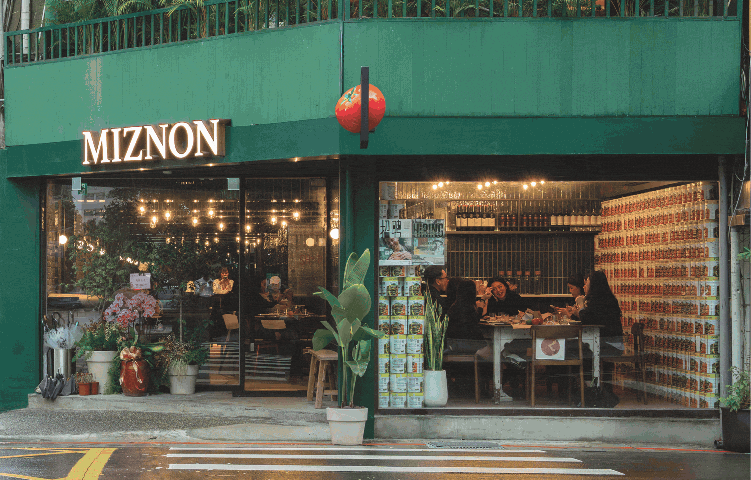 A green storefront restaurant named MIZNON with large glass windows showing people dining inside, decorated with potted plants outside, and a tomato logo above the entrance.