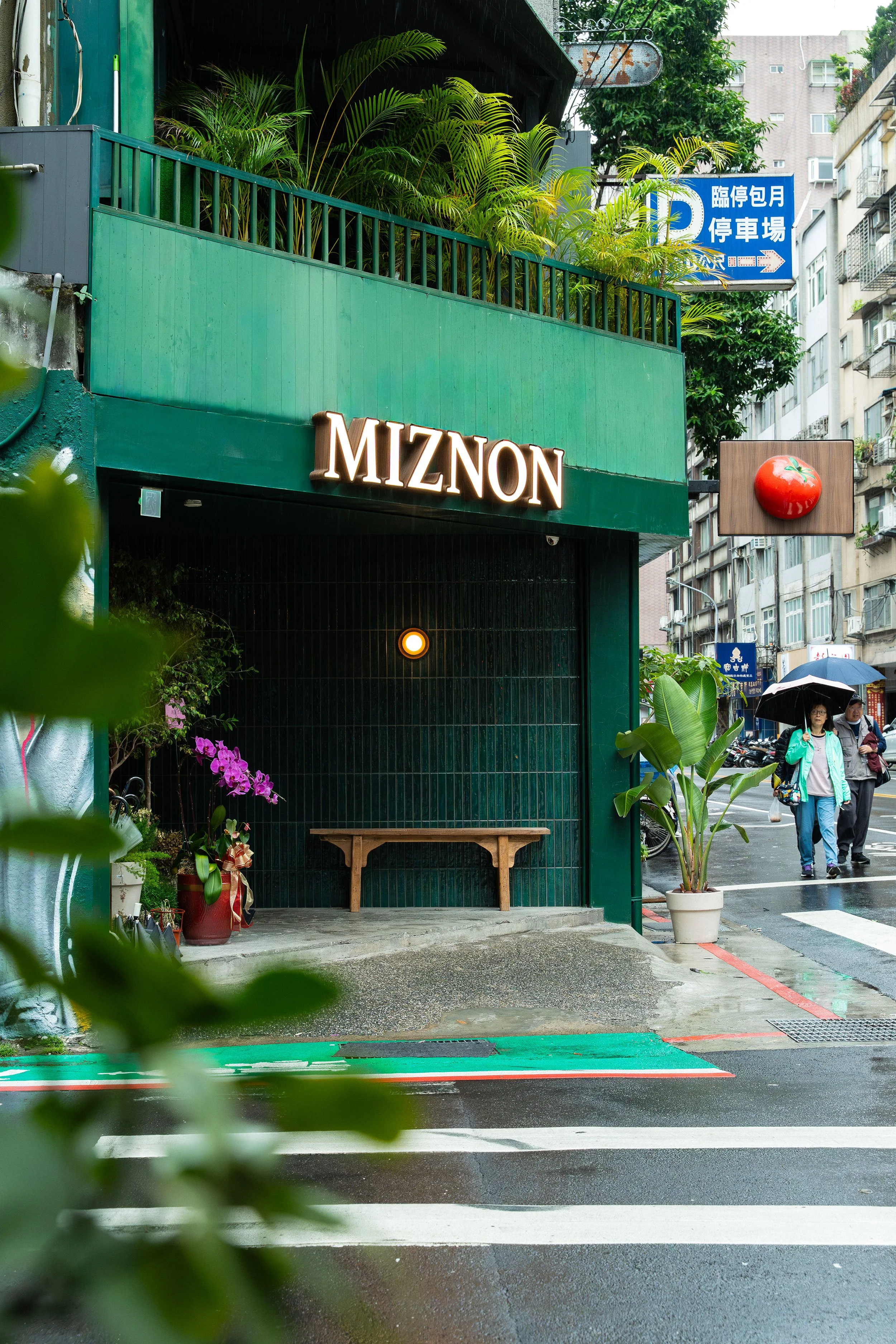 A storefront with a green facade and the sign 'MIZNON'. There are potted plants and flowers near the entrance, a wooden bench, and a green bench on the sidewalk. People are walking with umbrellas on a rainy day.