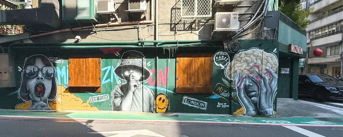 Colorful mural featuring three stylized portraits of women with exaggerated features, including large sunglasses, a person with a finger over lips, and a face with hands covering eyes, set against a green background with city names and street signs.