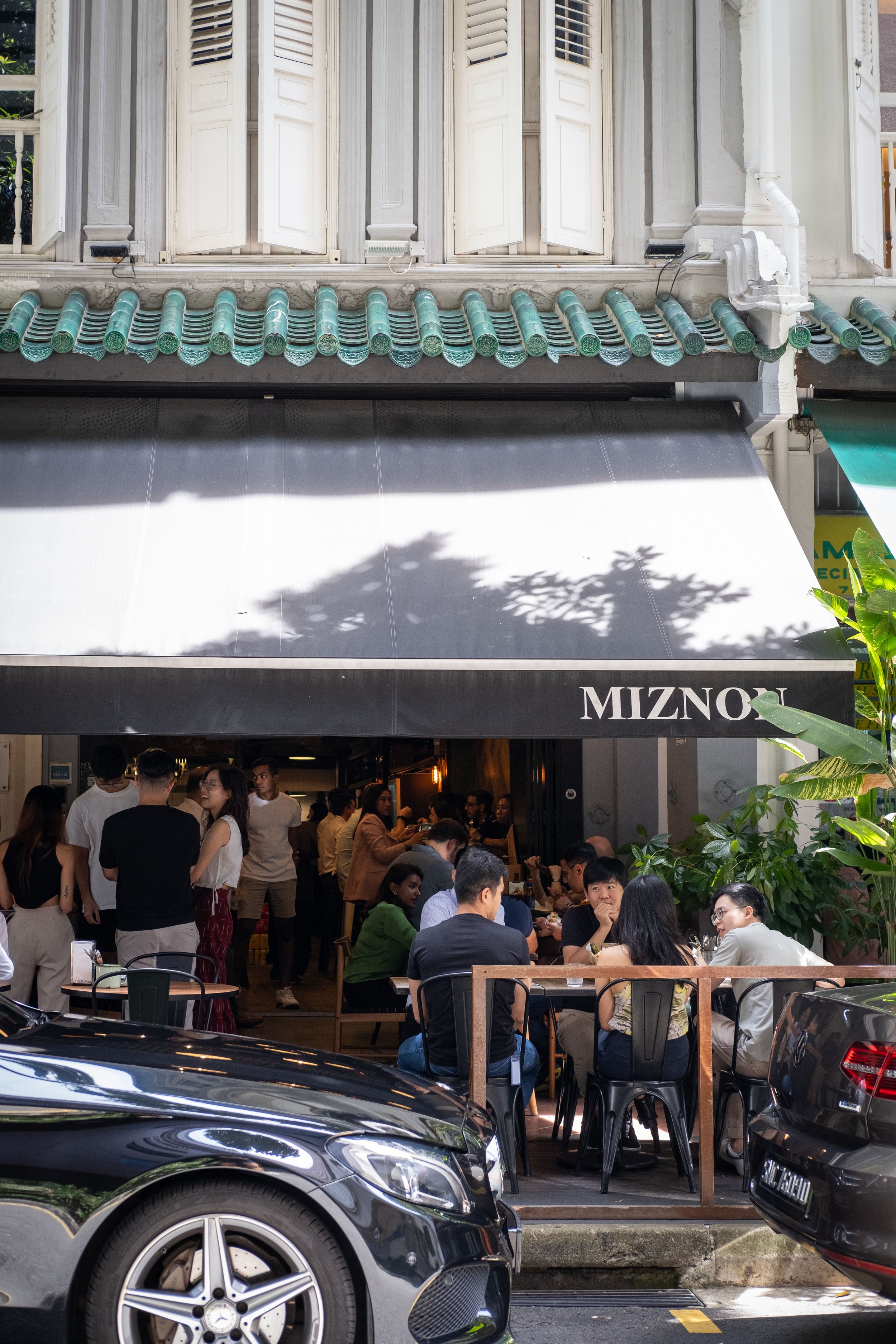 Crowded outdoor dining area in front of Mizno restaurant with people sitting at tables, cars parked nearby, and a gray awning with the restaurant name.