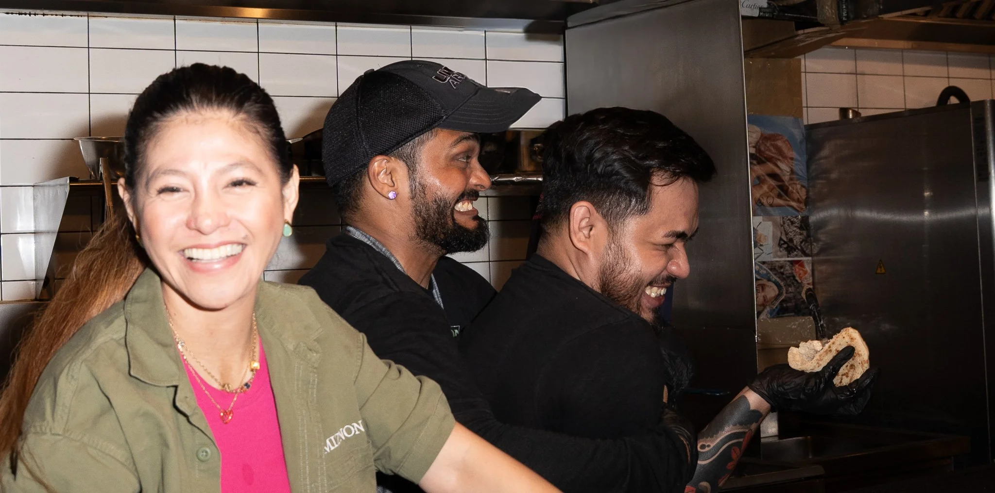 Four people smiling and working behind a counter in a kitchen. A woman with long hair and a green jacket in the foreground, three men in black shirts behind her, one wearing a cap, all smiling and engaged in food preparation.