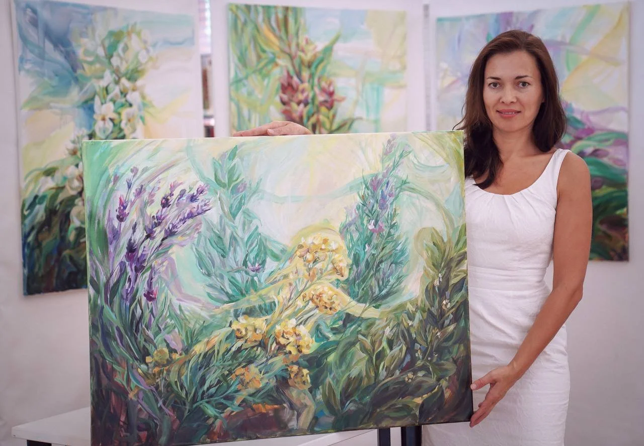 A woman in a white dress holding an abstract floral painting at an art gallery, with other colorful floral artworks hanging on the wall behind her.