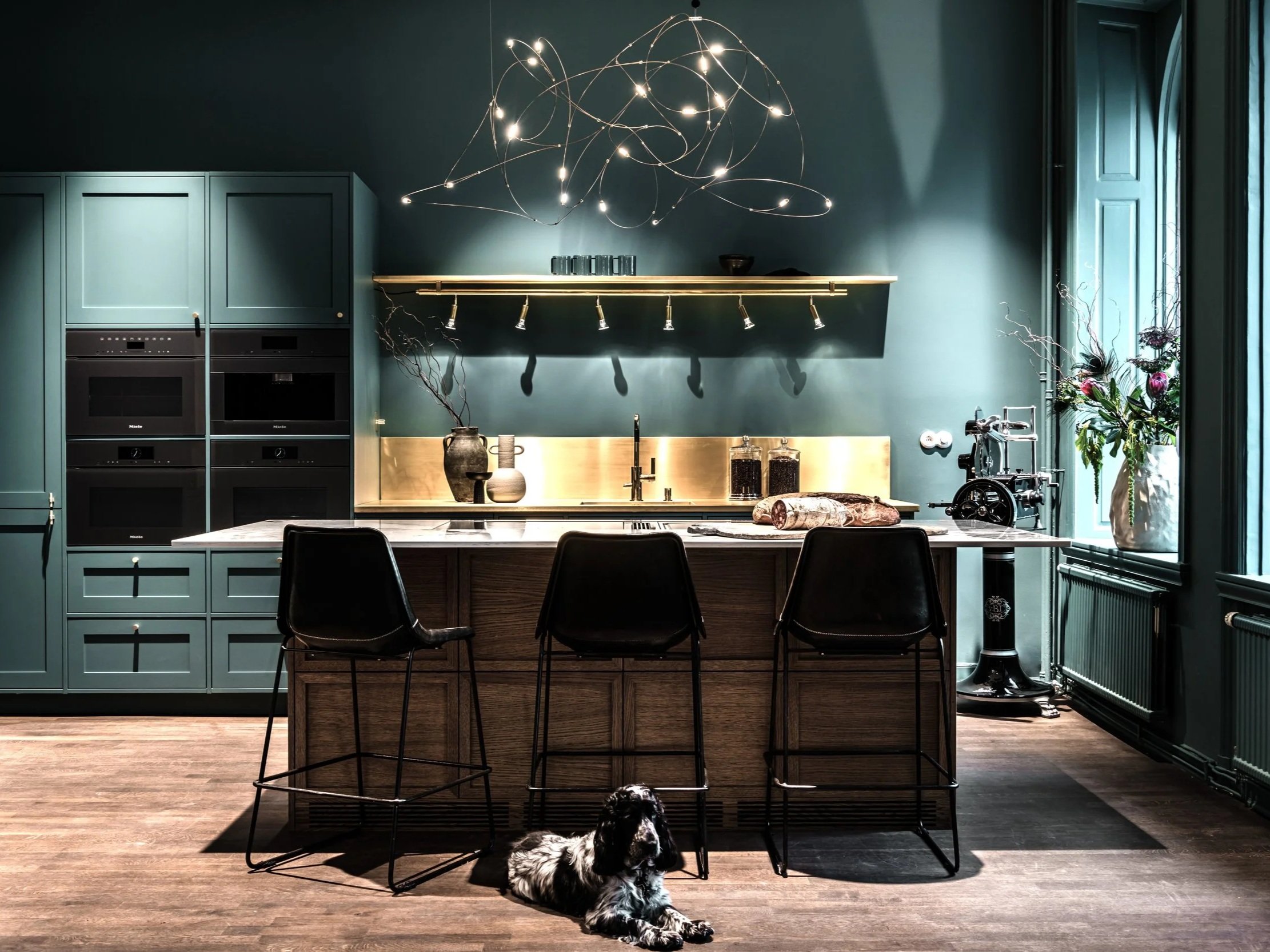 Modern kökskök med mörkgröna skåp, ett köksö med tre svarta barstolar, ett stjärnkroneliknande ljus ovanför, och en hund som ligger på golvet framför barstolarna.