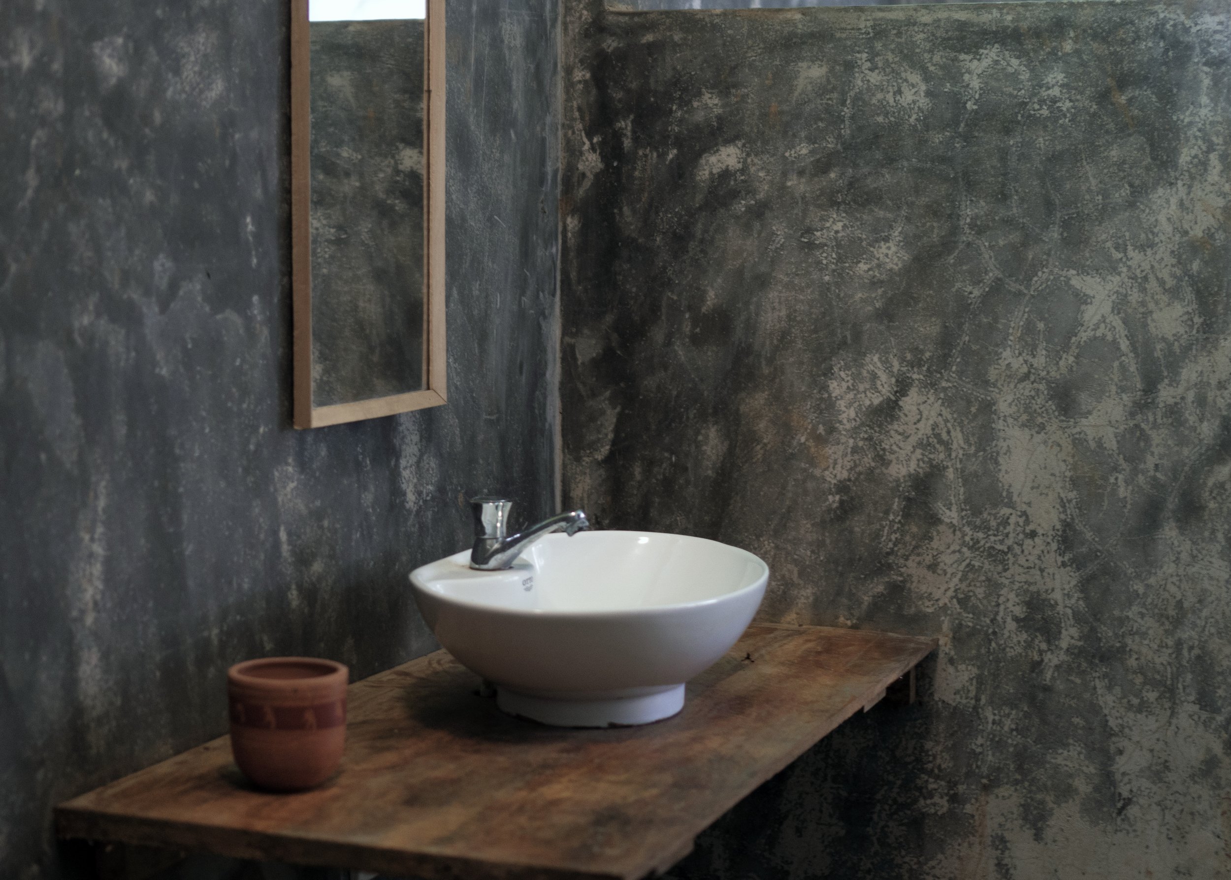 Modern bathroom with a white bowl sink on a wooden countertop, a small ceramic pot, and a rectangular mirror on a textured concrete wall. Cozy bathroom in Sri Lanka